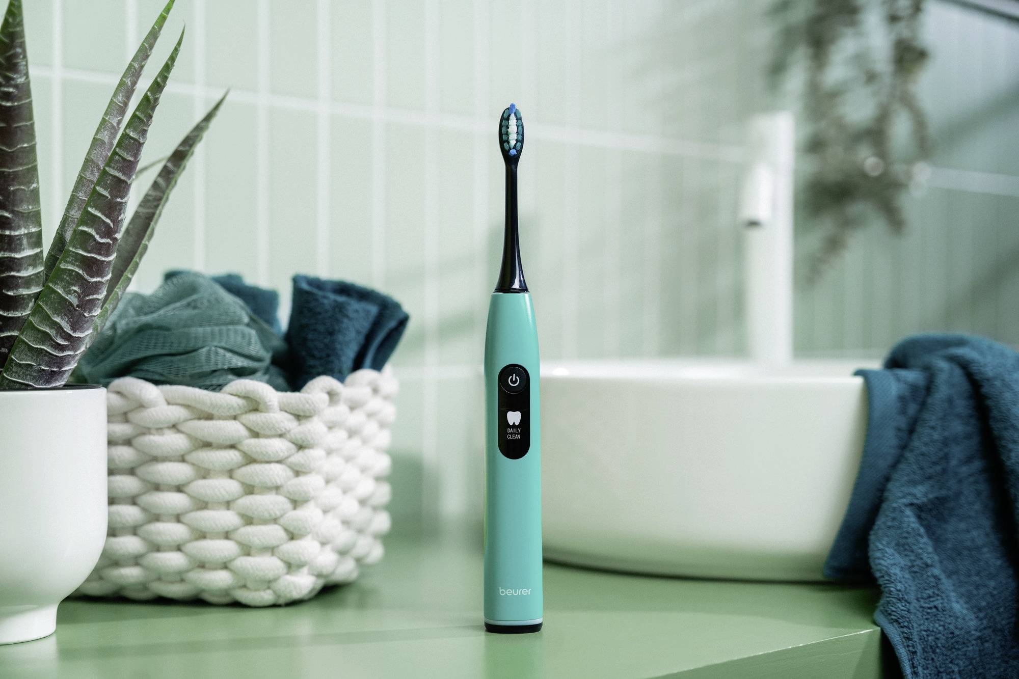 Eine türkisfarbene elektrische Zahnbürste steht aufrecht auf einer grünen Arbeitsplatte, neben einem geflochtenen Korb mit blauen Handtüchern, in einer Badezimmer-Umgebung.