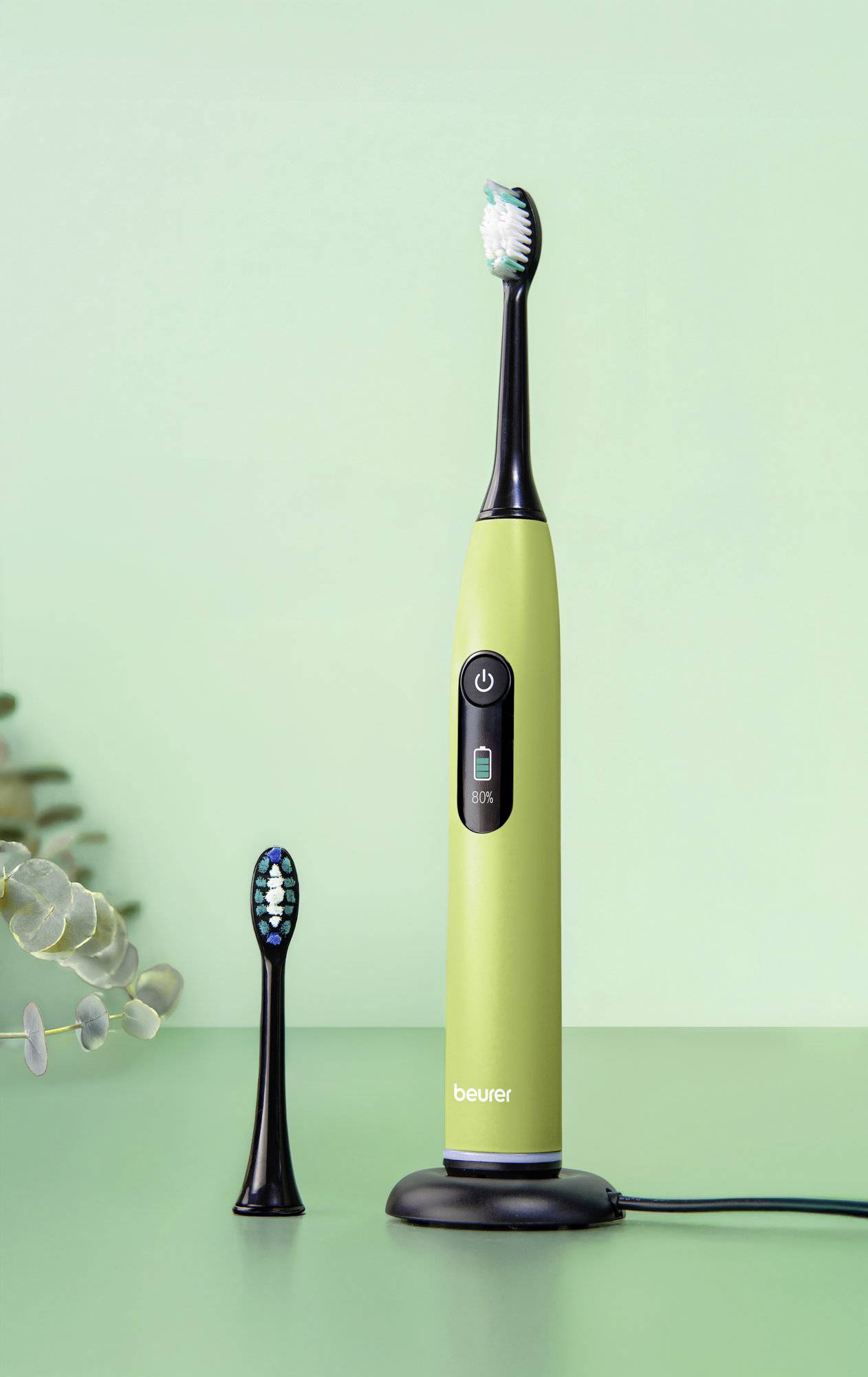Eine grüne elektrische Zahnbürste auf einer Ladestation, mit einem zusätzlichen Bürstenkopf in der Nähe, vor einem hellgrünen Hintergrund.