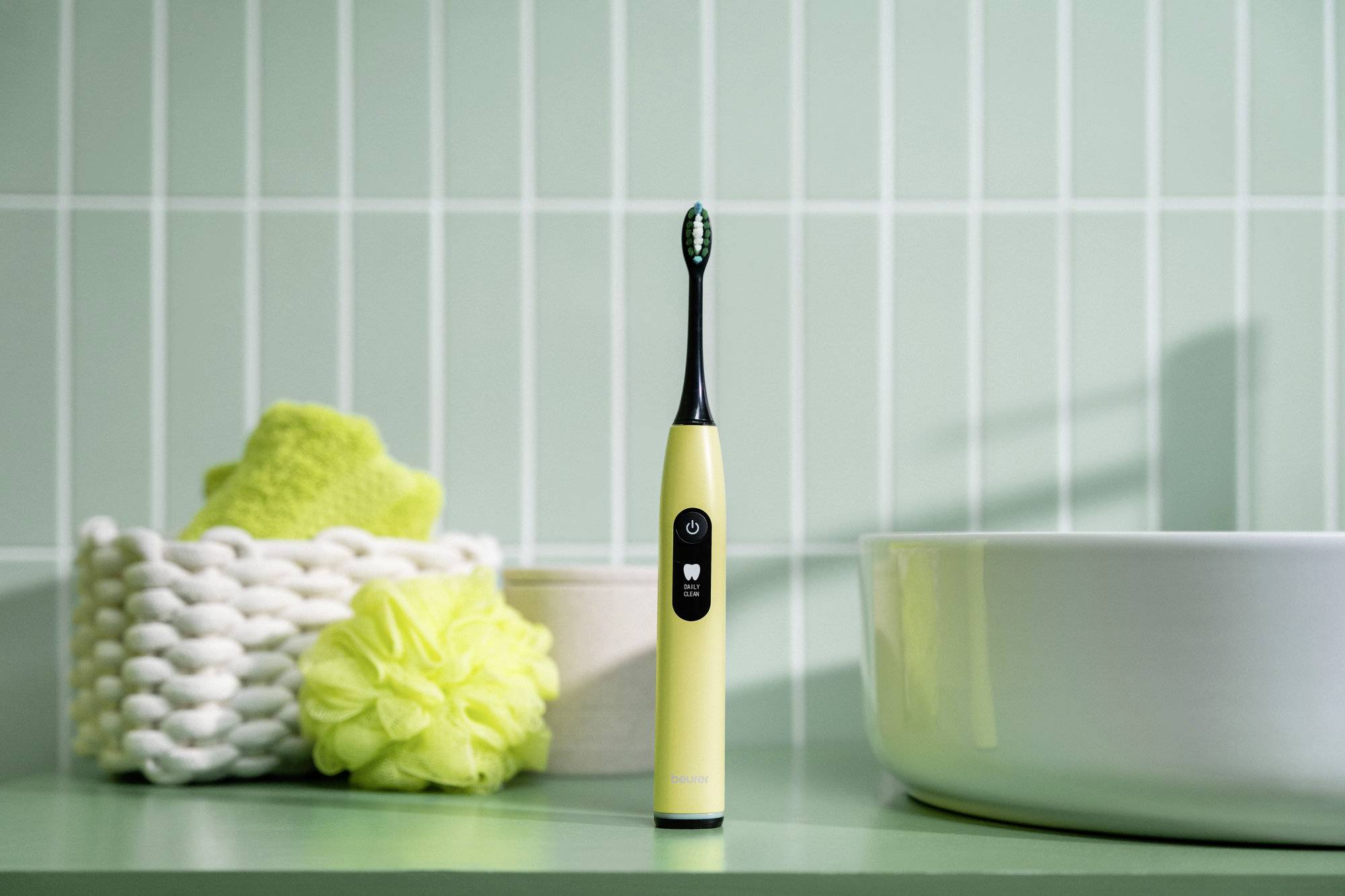 Eine gelbe elektrische Zahnbürste steht auf einem grünen Badezimmertresen. Links daneben befindet sich ein geflochtener Korb mit grünen Handtüchern und einem Badeschwamm.