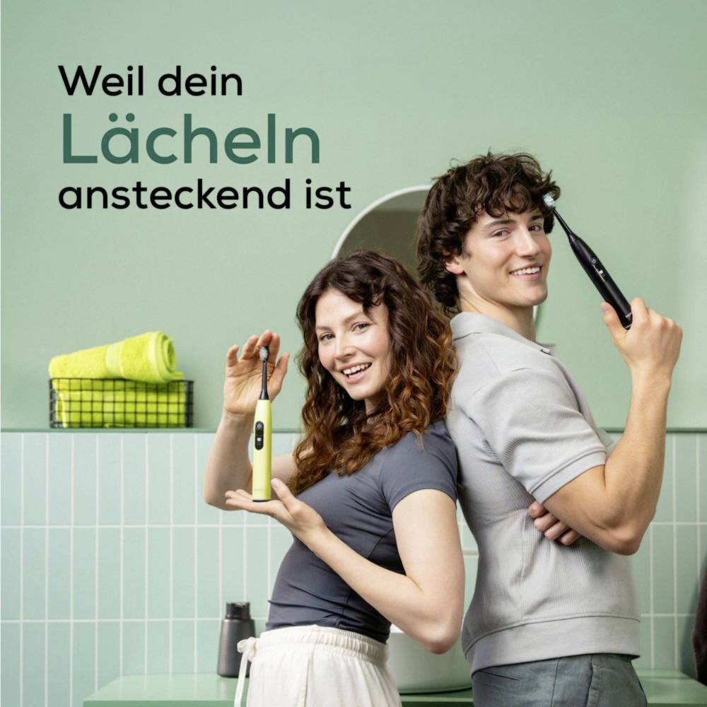 A smiling couple holding electric toothbrushes in a bathroom. Text reads 'Weil dein Lächeln ansteckend ist' ('Because your smile is contagious').<br><br>(Note: The text is already in German, so no translation is needed.)