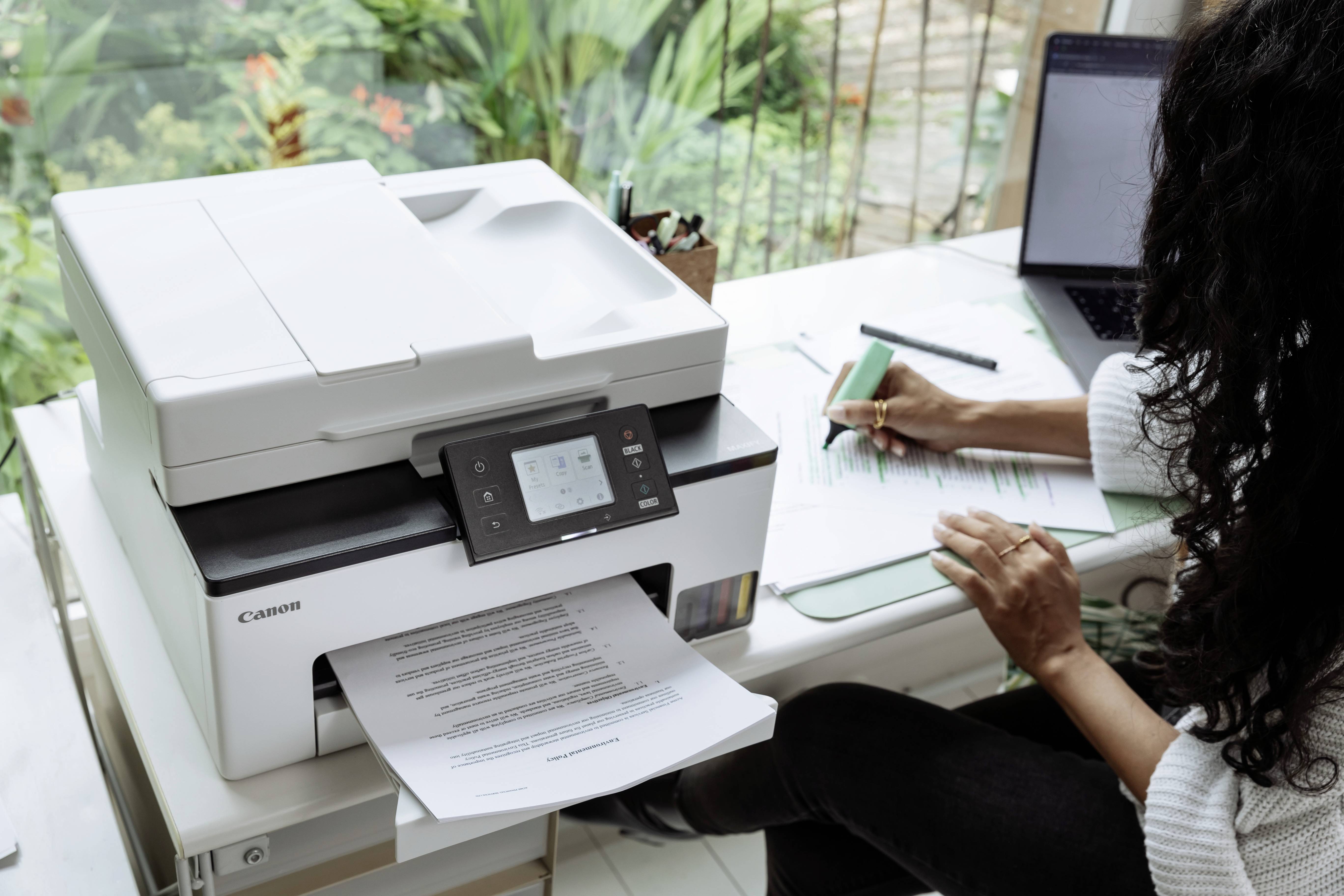 Eine Person sitzt an einem Schreibtisch in einem Heimarbeitsplatz und verwendet einen Drucker neben einem Laptop. Der Drucker druckt gerade ein Dokument aus.