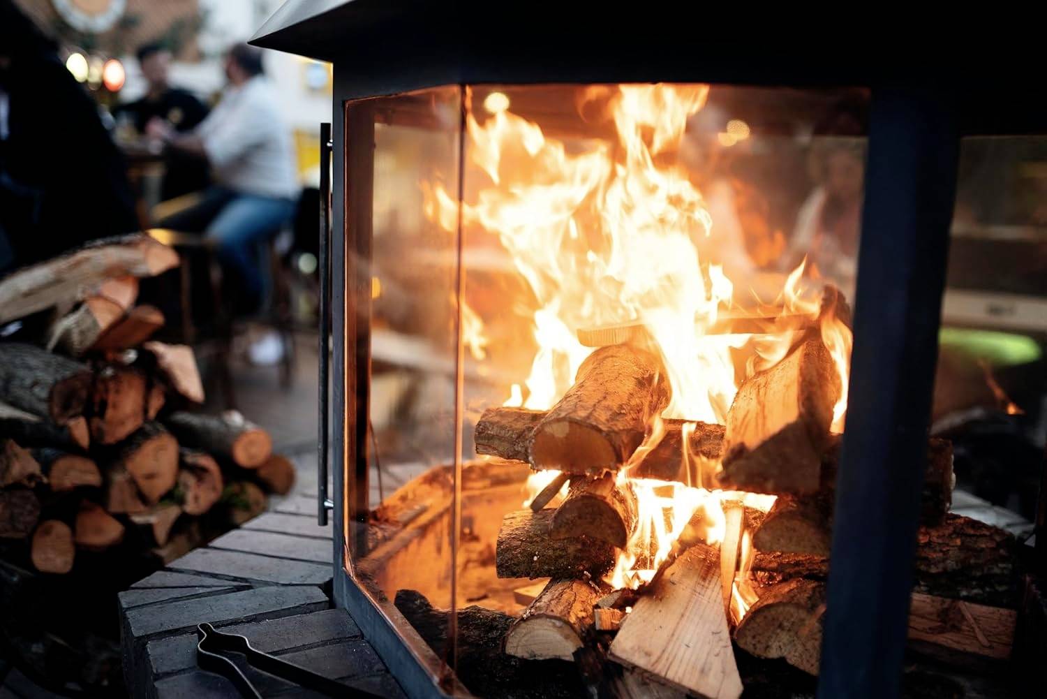 Ein warmes, glühendes Feuer brennt in einem Metallkamin im Freien, mit gestapelten Holzscheiten in der Nähe. Menschen sind im Hintergrund unscharf, was auf eine gemütliche Zusammenkunft hindeutet.