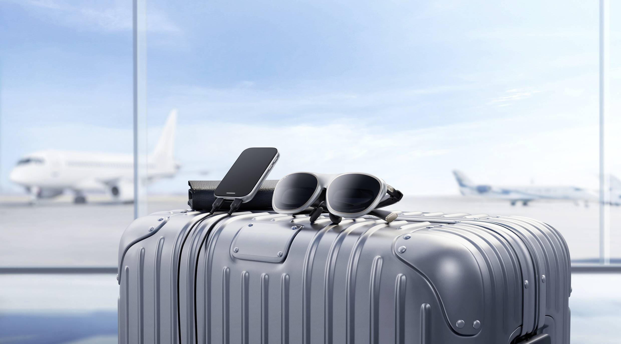 Ein silberner Koffer auf einem Flughafen mit einem Smartphone und einer Sonnenbrille obenauf. Zwei Flugzeuge sind durch große Fenster im Hintergrund sichtbar.