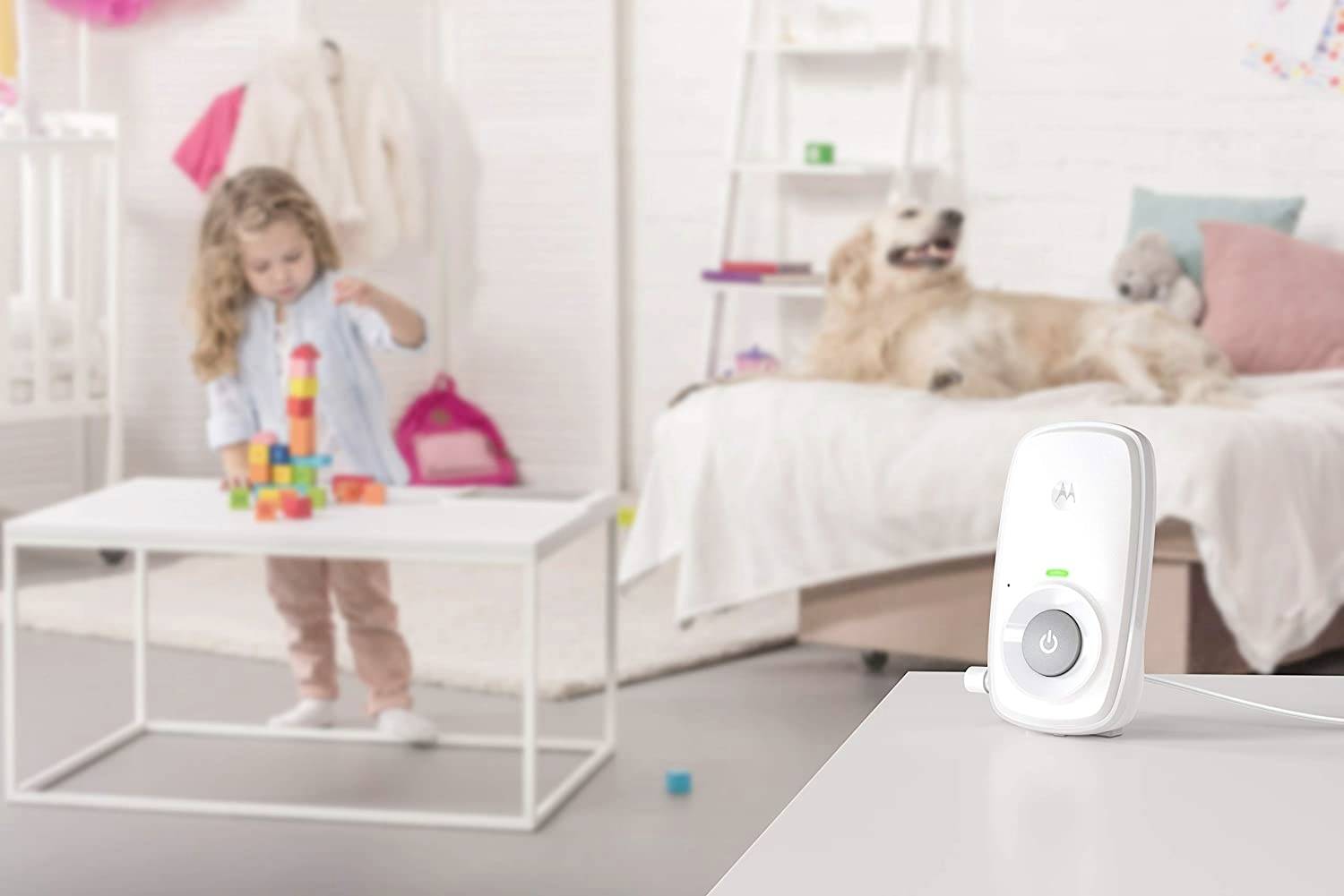 Ein Babyphone steht im Vordergrund auf einem Tisch. Im Hintergrund spielt ein Kind mit Bauklötzen, und ein Hund ruht auf einem Bett.