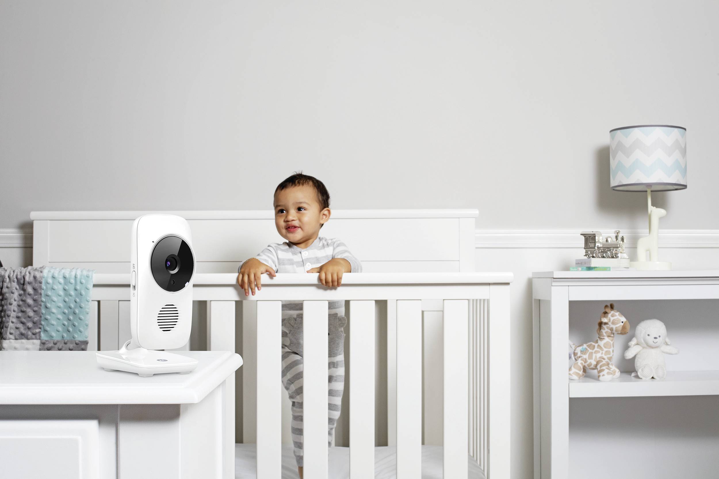 Ein Kleinkind steht in einem weißen Gitterbett und schaut über die Seitenlehne. Ein Babyphone ist in der Nähe positioniert, und im Hintergrund befindet sich ein Regal mit Spielzeugen und einer Lampe.