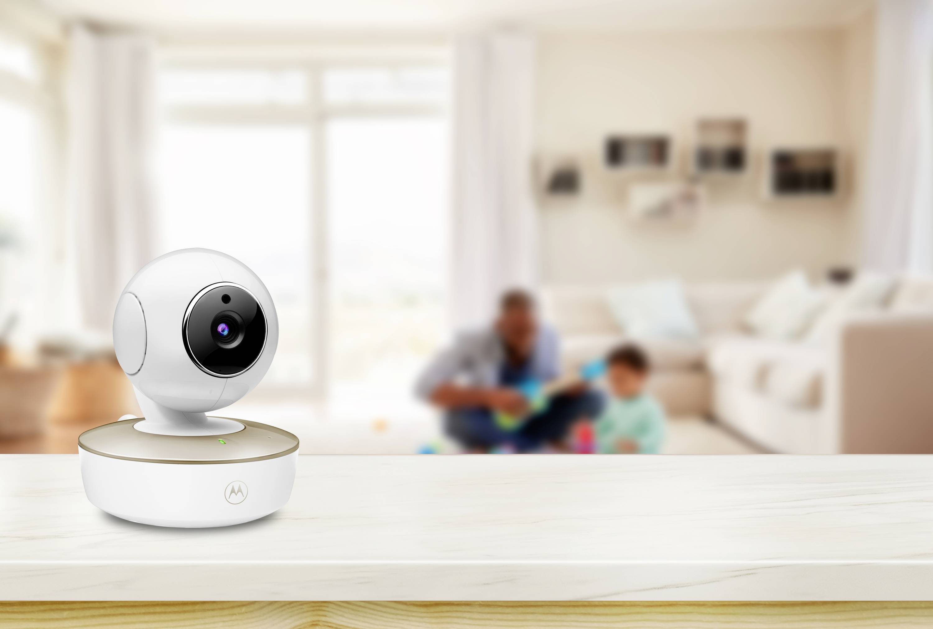 Eine moderne Indoor-Sicherheitskamera steht auf einer Holzoberfläche in einem hellen Wohnzimmer. Im unscharfen Hintergrund spielen ein Erwachsener und ein Kind.