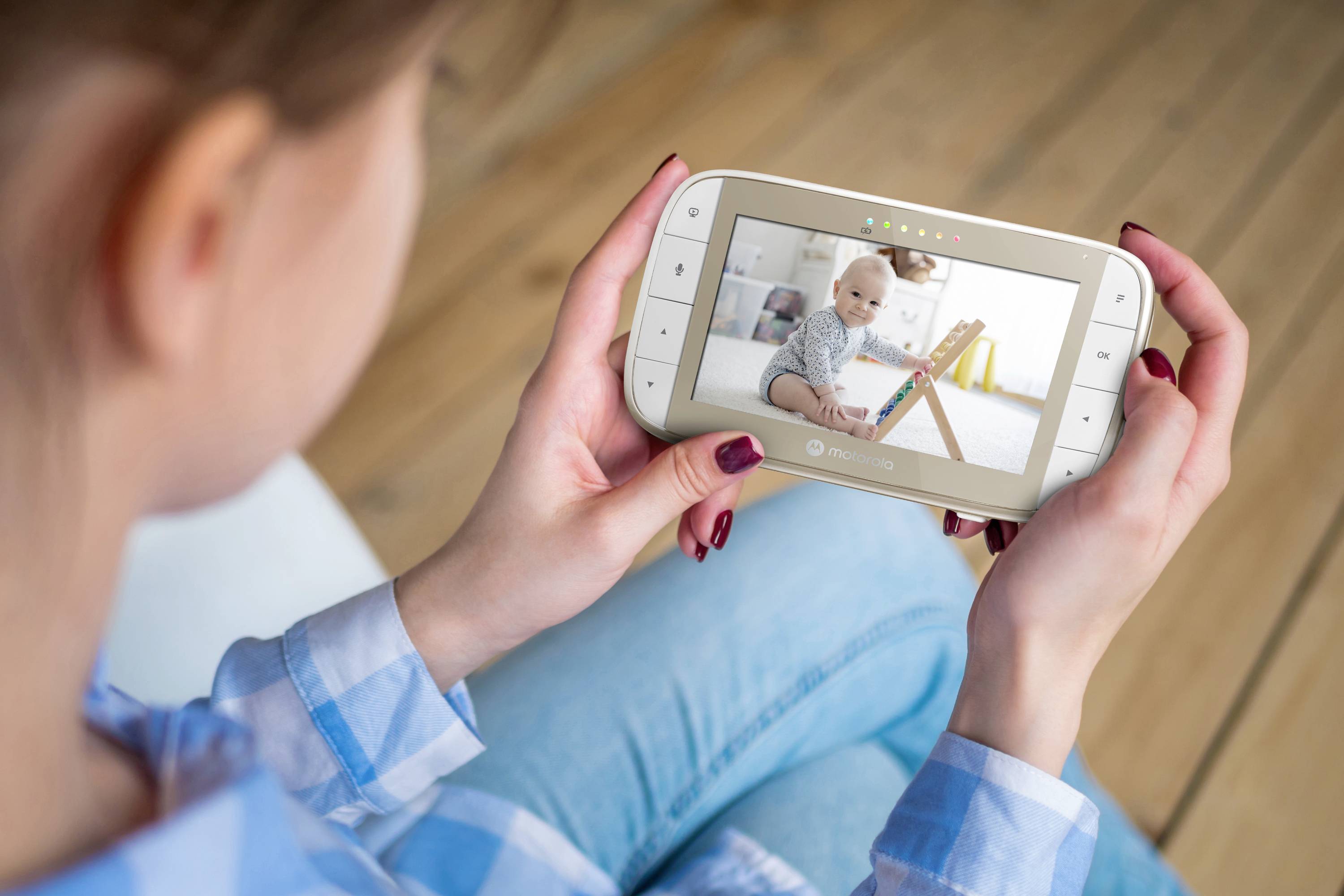 Eine Frau hält einen Babyphone, auf dem ein Baby auf dem Boden spielt und mit einem kleinen Holzspielzeug beschäftigt ist.