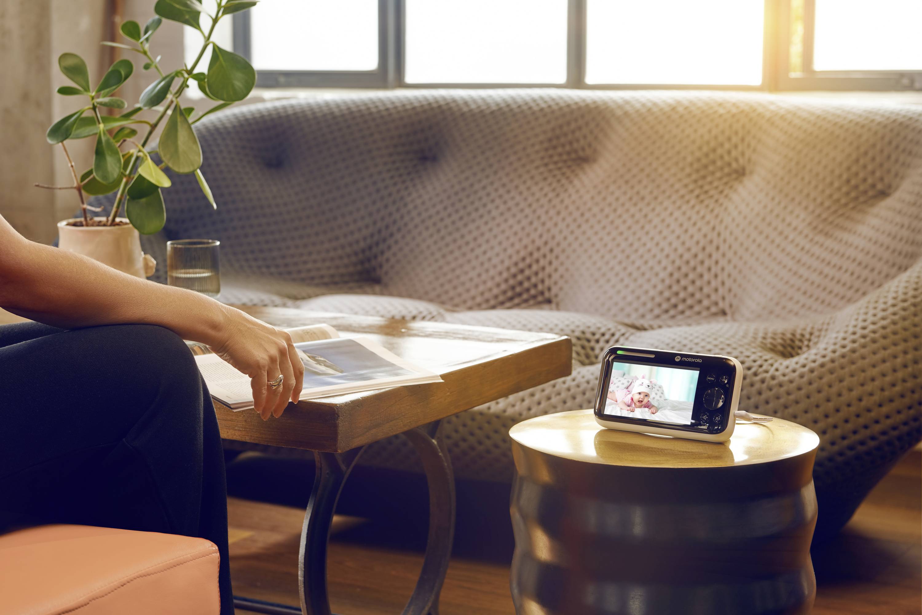 Eine Person liest an einem Tisch in einem sonnendurchfluteten Wohnzimmer. Auf dem runden Tisch zeigt ein Smartphone einen Videoanruf mit einem Kind.