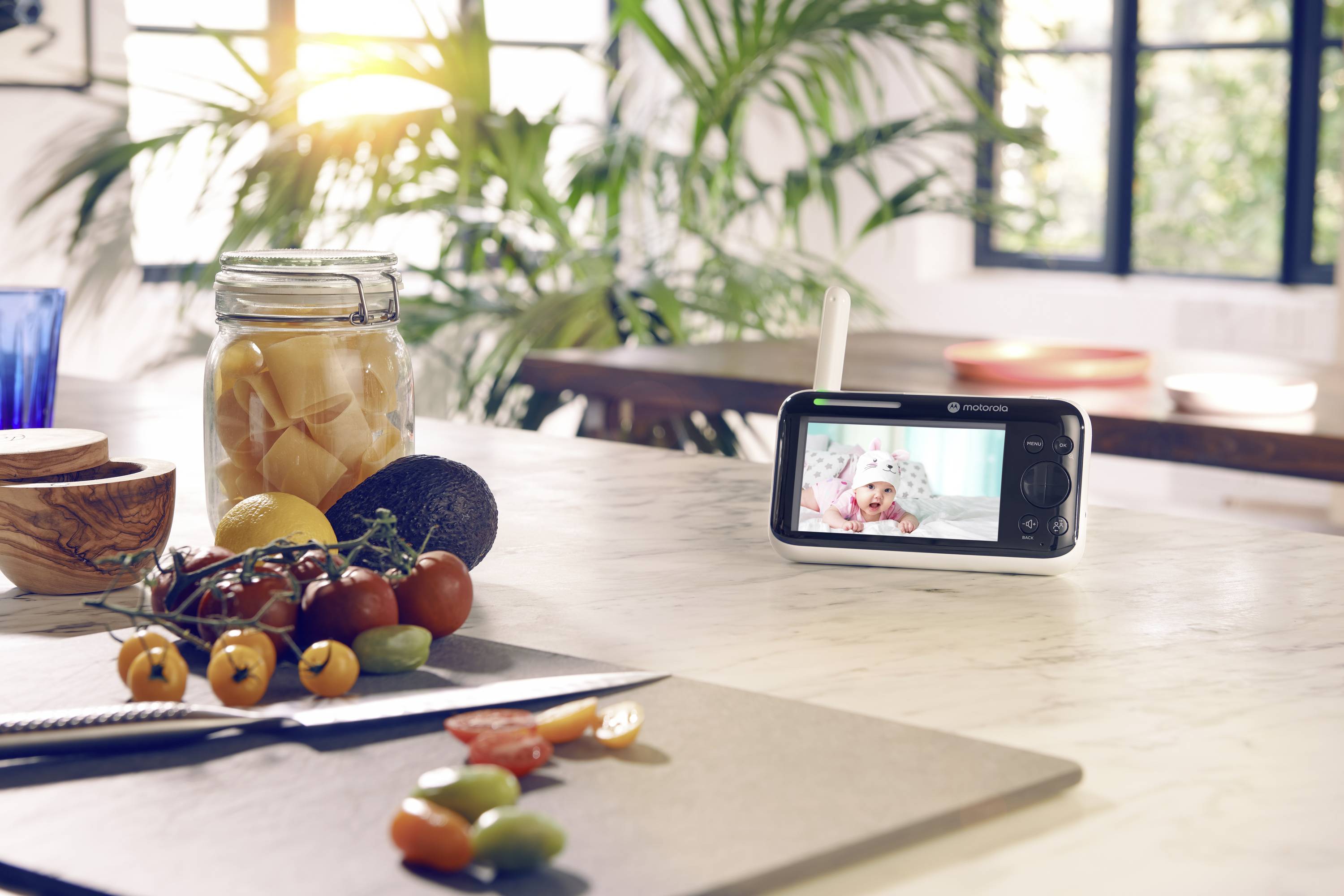 Eine Küchenarbeitsplatte mit einem Schneidebrett, Tomaten, einem Einmachglas mit Früchten und einem Babyphone, das ein spielendes Baby zeigt, vor dem Hintergrund eines sonnendurchfluteten Raumes.