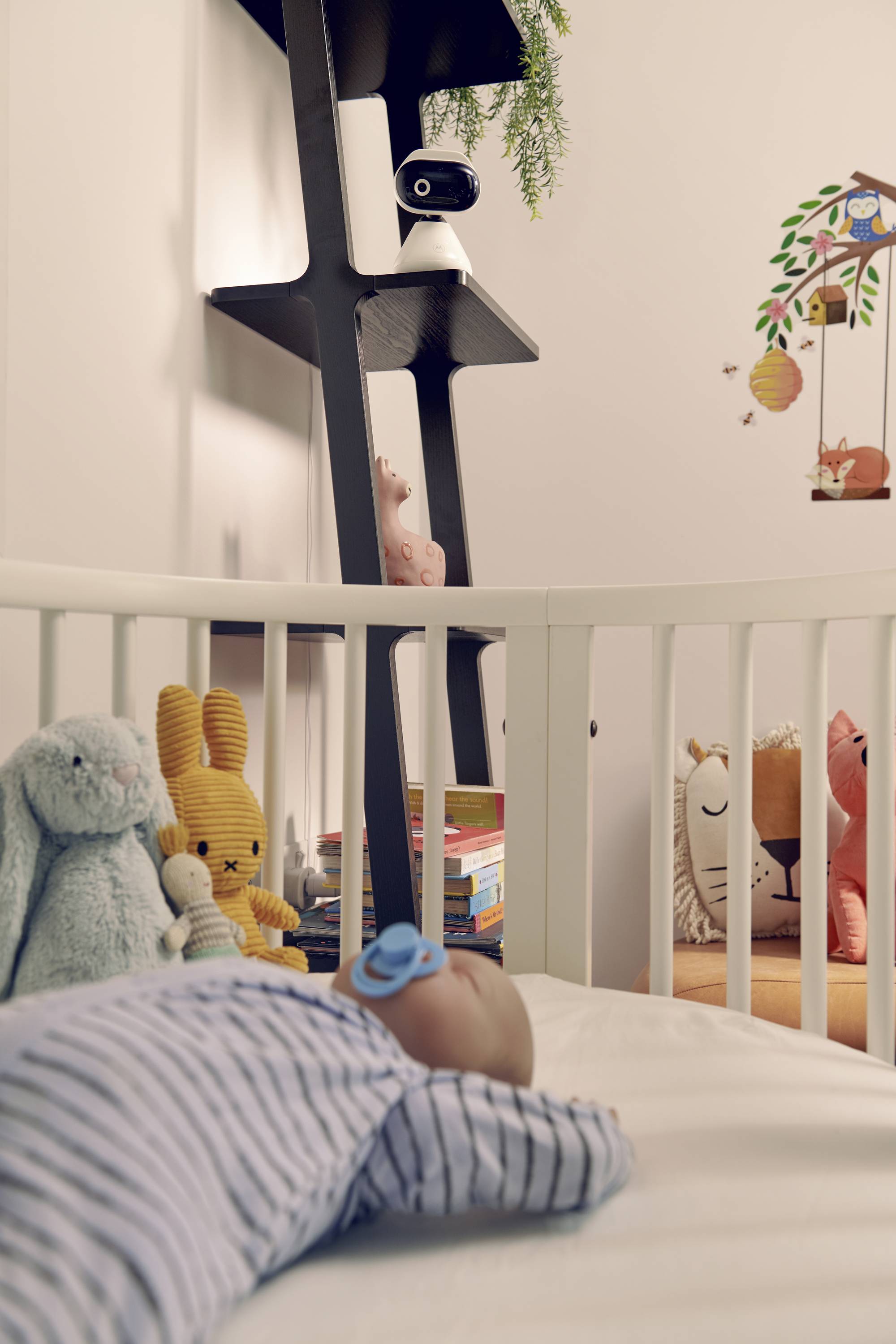 Ein Baby schläft in einer Wiege, umgeben von Plüschtieren. Ein schwarzes Regal mit Büchern und einer Kamera überblickt die Szene. An der Wand sind Tieraufkleber angebracht.