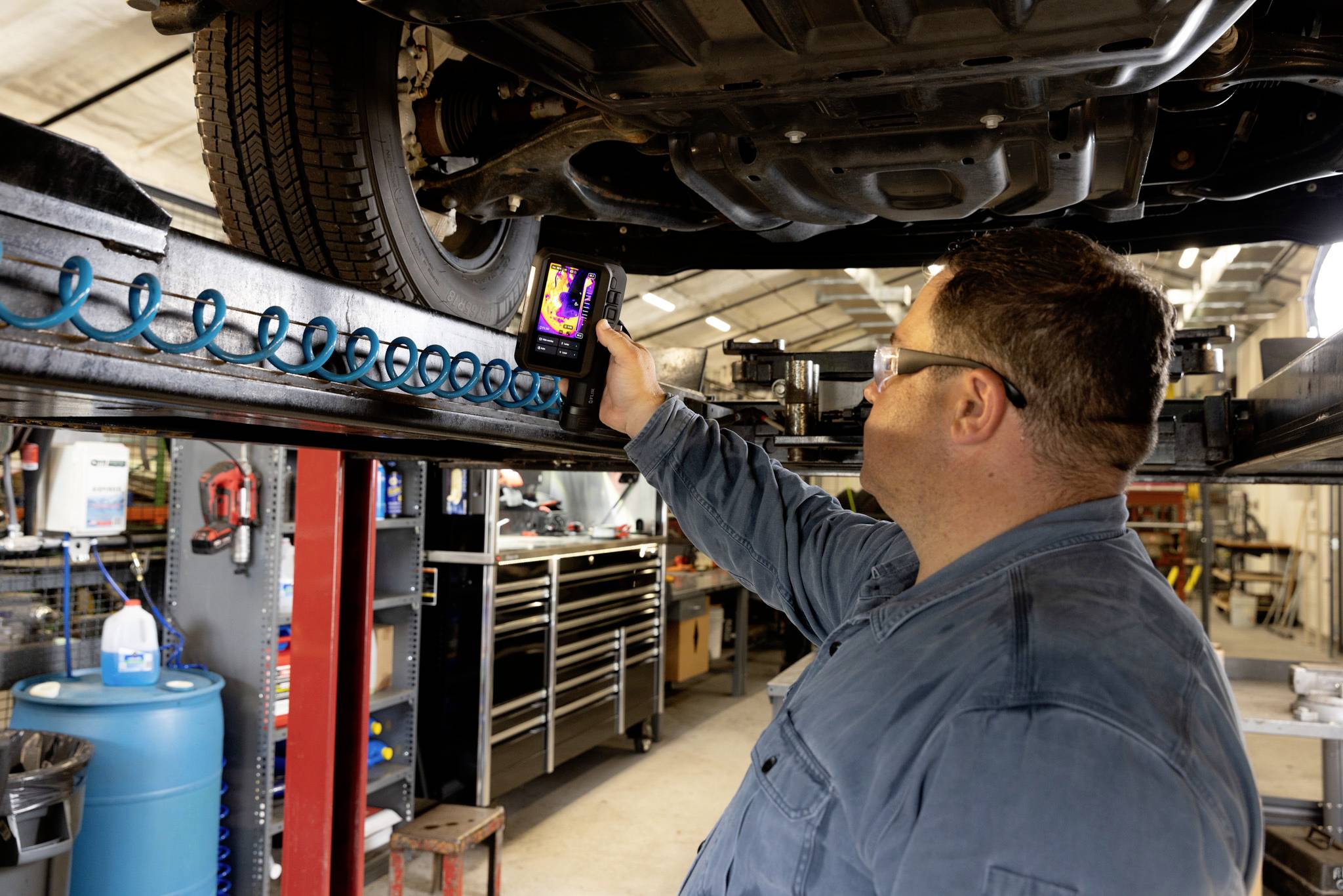 Ein Mechaniker in einer Werkstatt verwendet ein Wärmebildgerät zur Untersuchung der Fahrzeugunterkonstruktion. Im Hintergrund sind verschiedene Werkzeuge zu sehen.