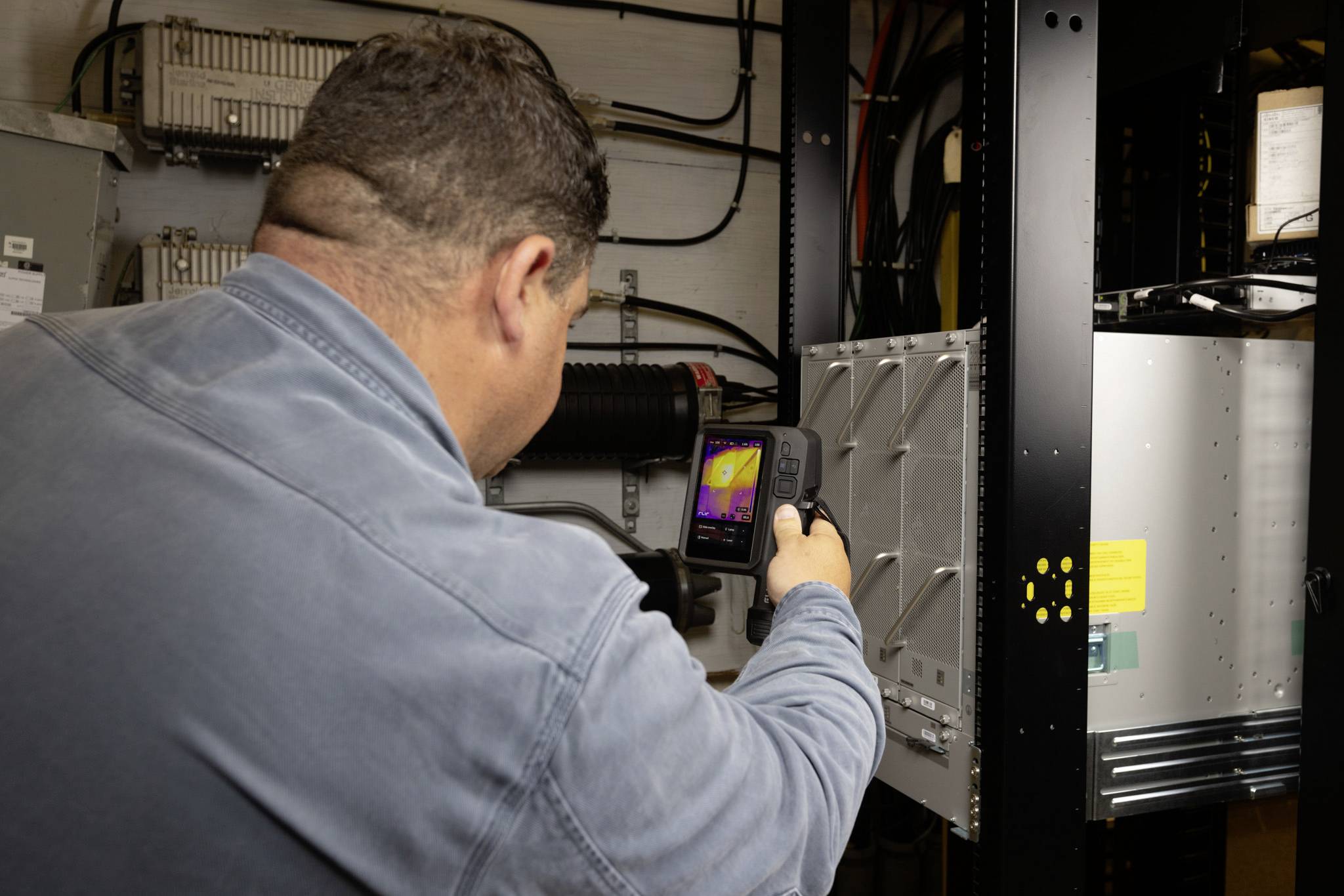 Eine Person verwendet eine Wärmebildkamera, um elektronische Geräte in einem Serverraum zu untersuchen und dabei Hotspots oder Fehlfunktionen zu überprüfen.