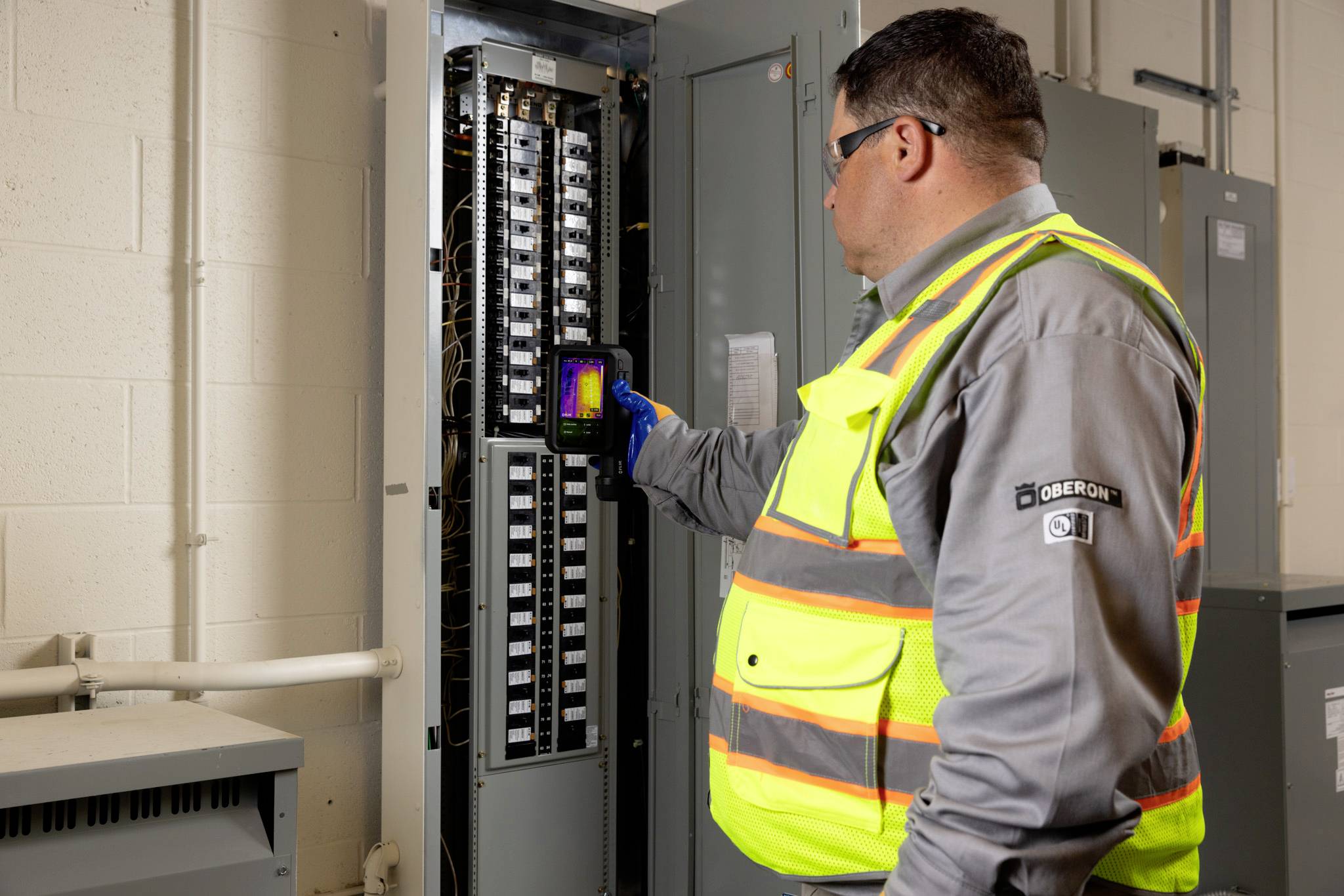 Ein Techniker in einer Sicherheitsweste verwendet eine tragbare Wärmebildkamera, um eine offene Elektrische Tafel zu inspizieren und dabei Sicherheit und einwandfreie Funktion zu gewährleisten.