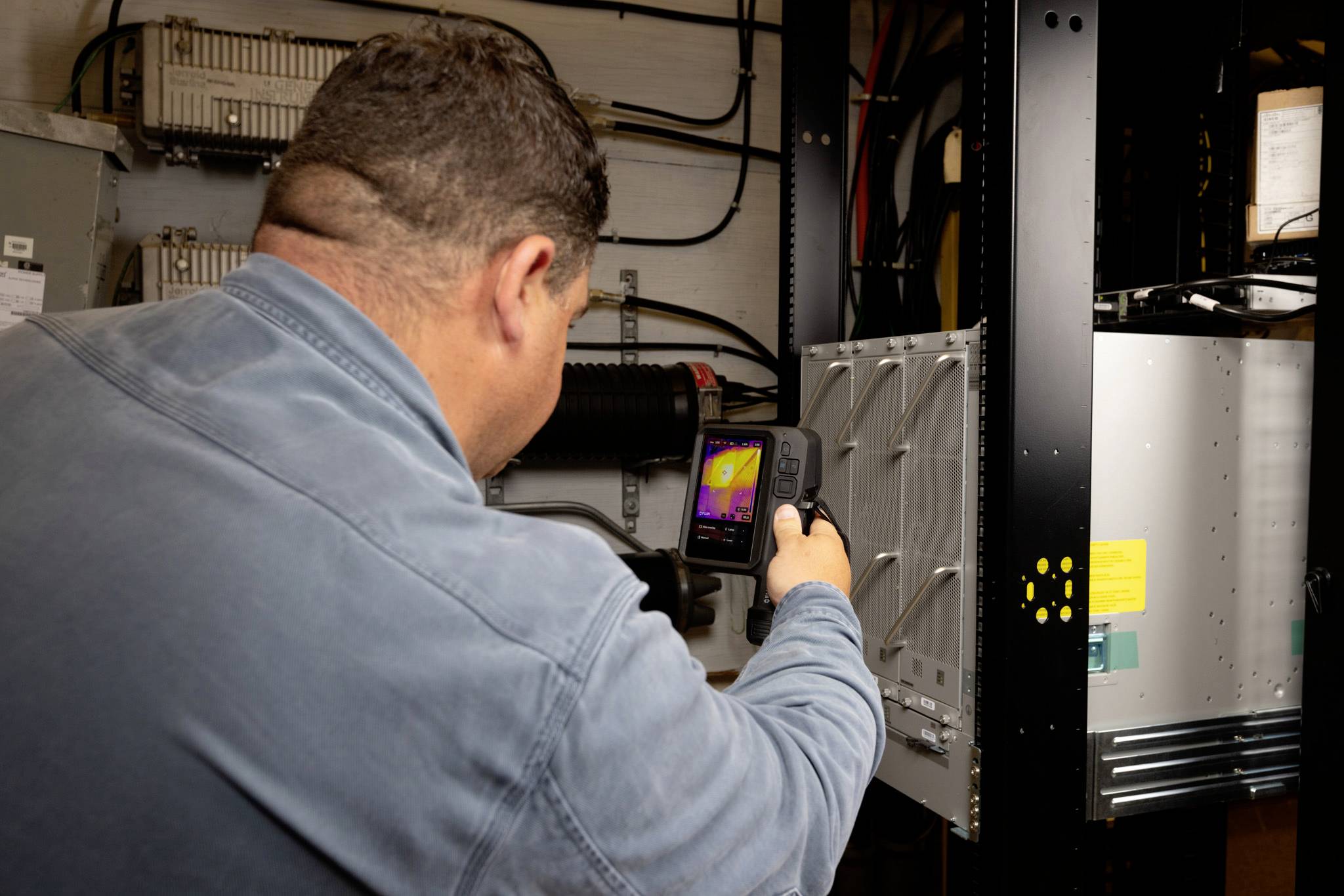 Eine Person nutzt ein Wärmebildgerät zur Inspektion von Elektrotafeln in einer industriellen Umgebung und erfasst die Wärmeverteilung auf dem Bildschirm.