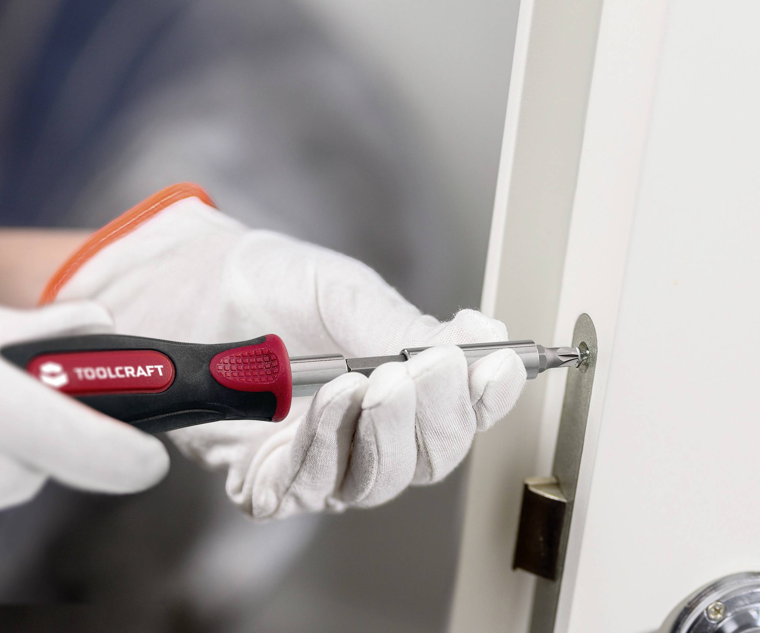 Eine Person mit Handschuhen verwendet einen Schraubendreher, um eine Schraube an einer Türangel festzuziehen und zeigt dabei sicheren Werkzeugumgang.