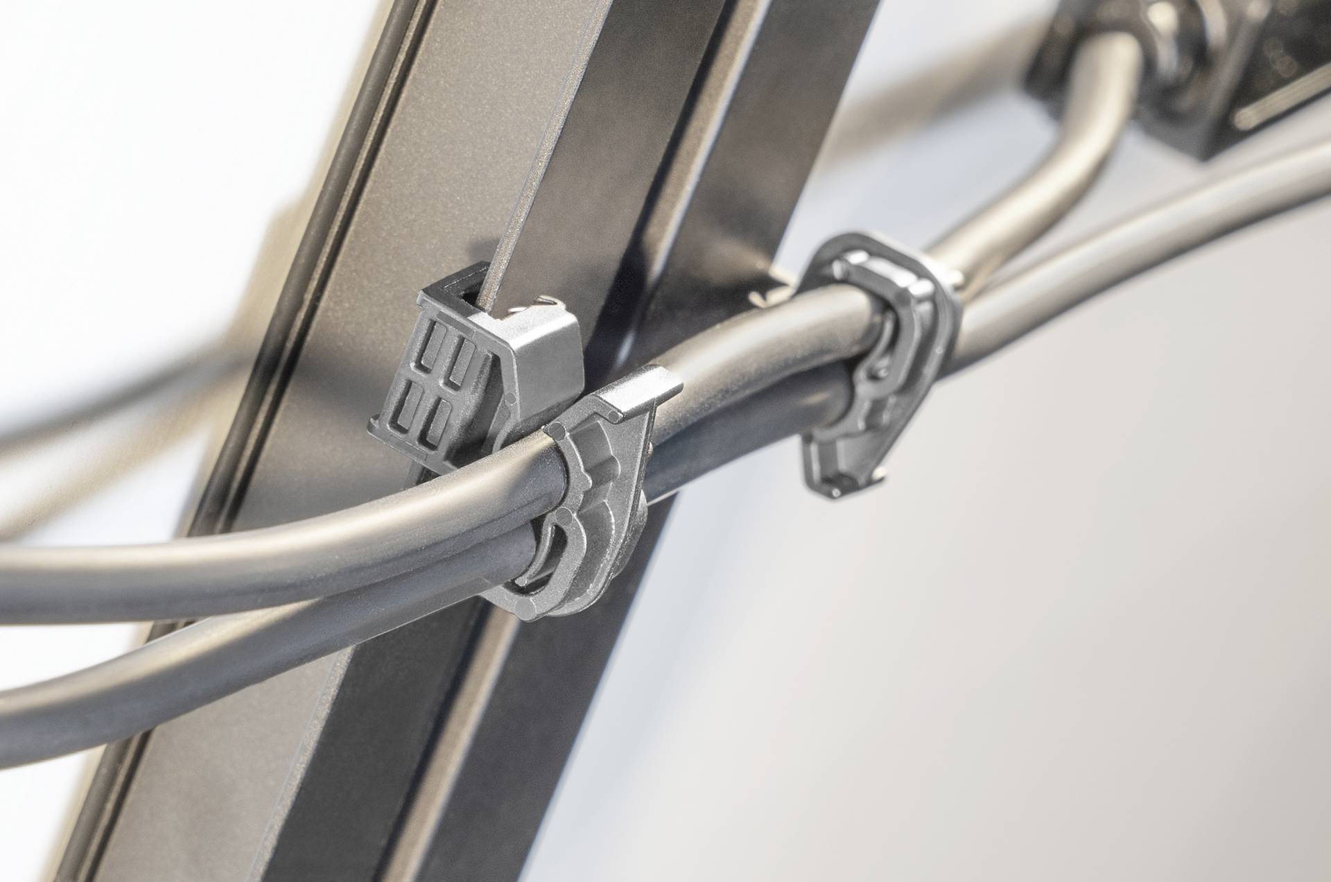 Nahaufnahme von zwei schwarzen Kabeln, die mit Kunststoffklammern sicher an einer vertikalen Metallschiene an einer hellgefärbten Wand befestigt sind.