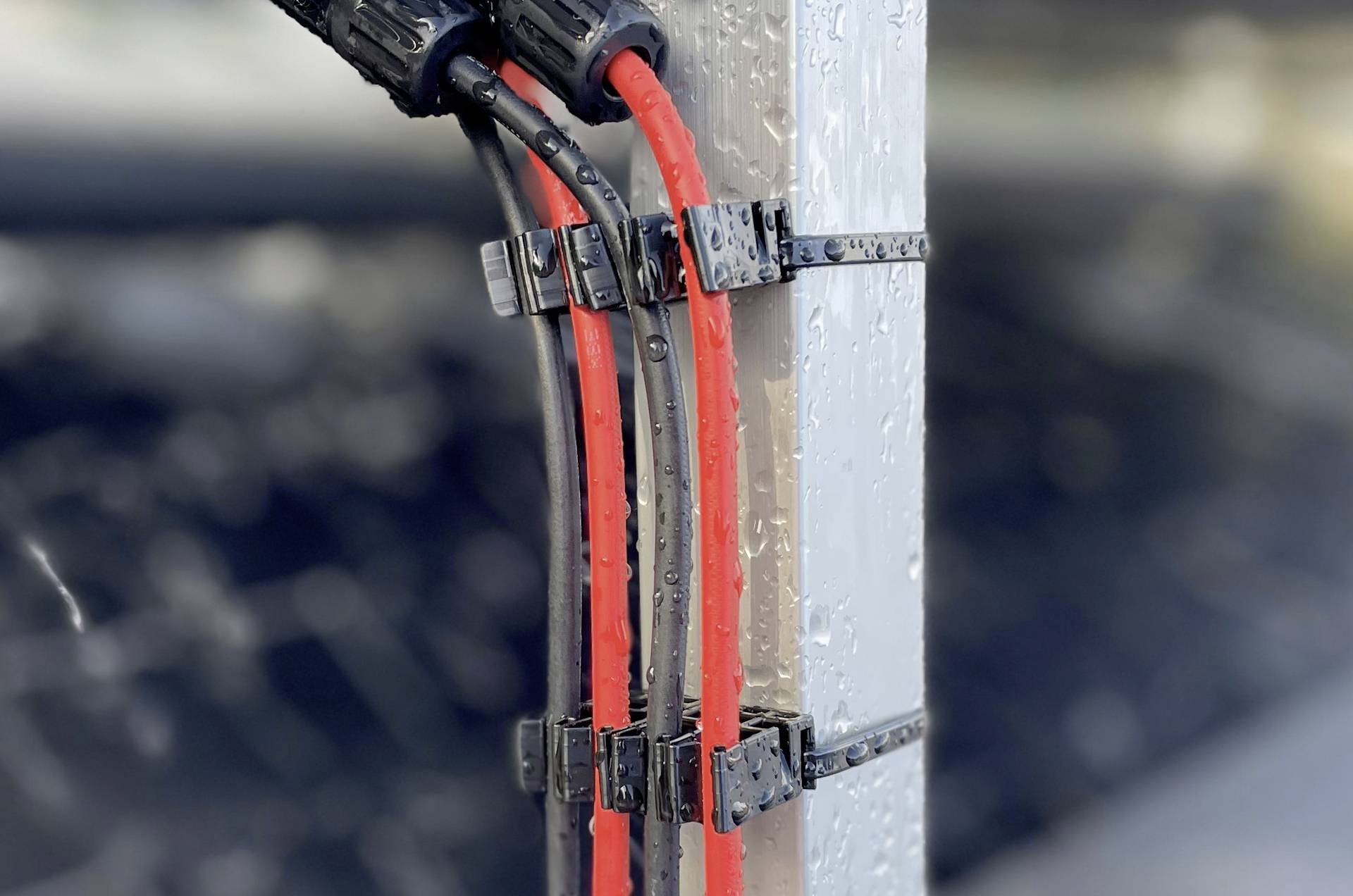 Rote und schwarze Kabel, die mit Kabelbindern an einem Metallmast befestigt sind, vor einem unscharfen Hintergrund. Die Kabel wirken feucht und deuten auf einen Einsatz im Freien hin.