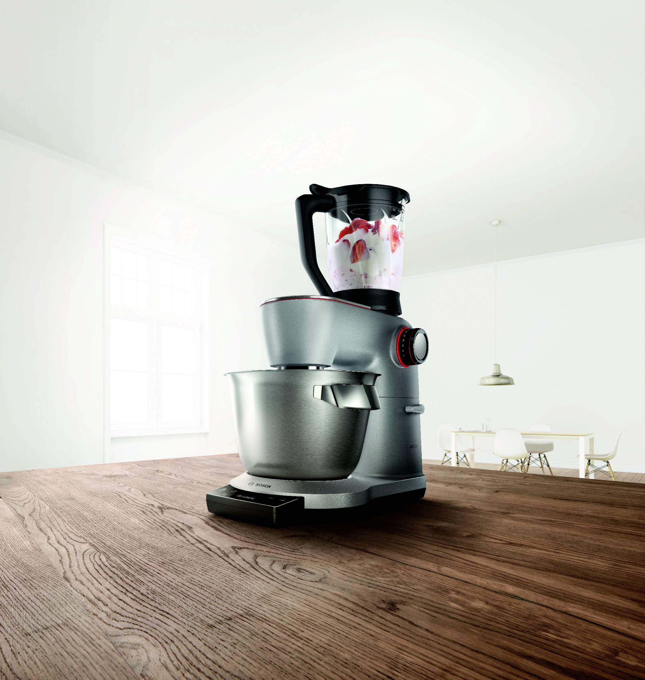 Ein silberner Küchenstandmixer mit Blenderaufsatz auf einer Holzarbeitsplatte in einer hellen, modernen Küche mit weißen Wänden und einem Fenster.