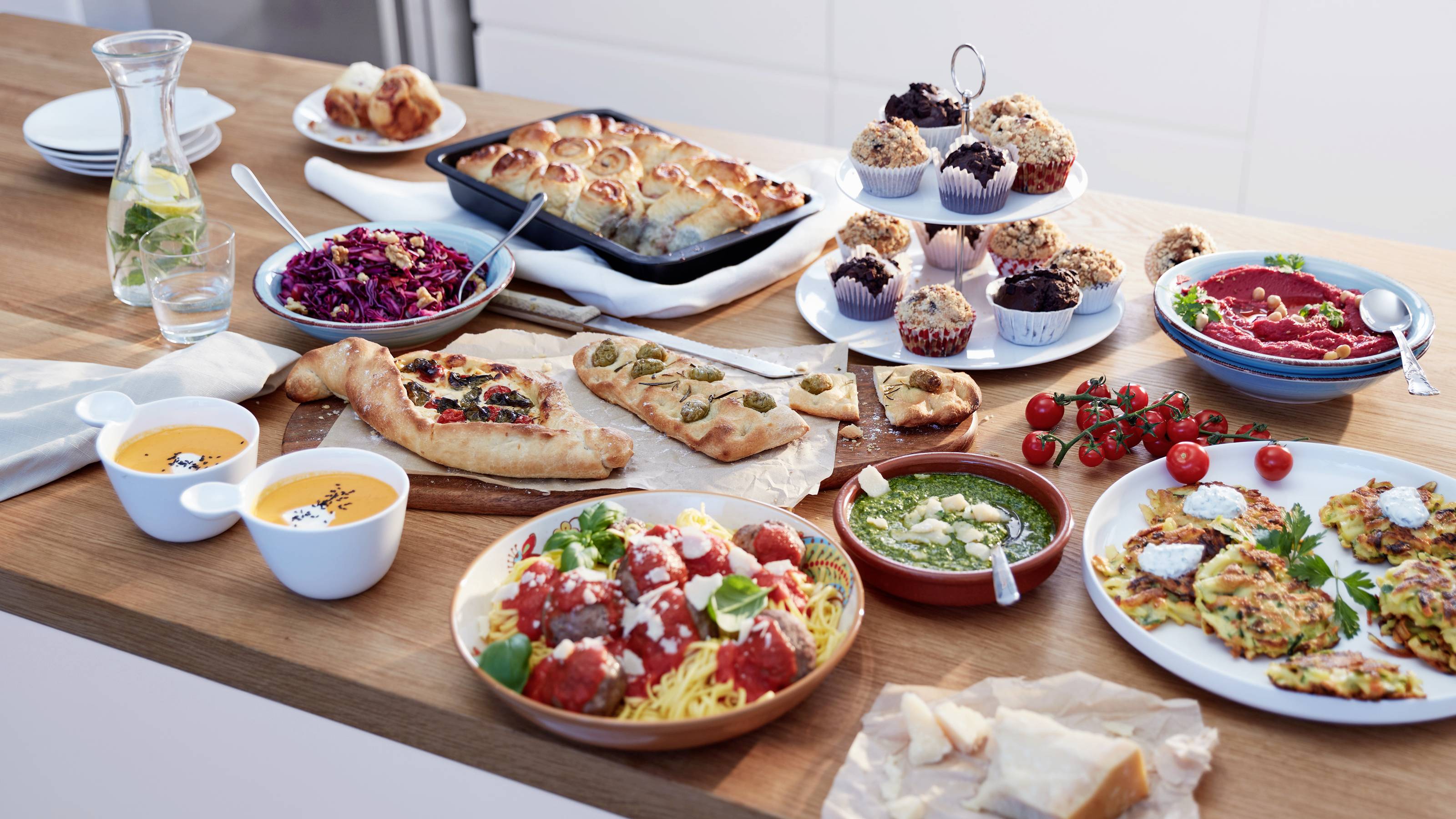 Ein Holztisch, der mit vielfältigen Gerichten bestückt ist, darunter Cupcakes, Salate, Suppen, Pasta mit Fleischbällchen und Brot, für ein Buffet angerichtet.