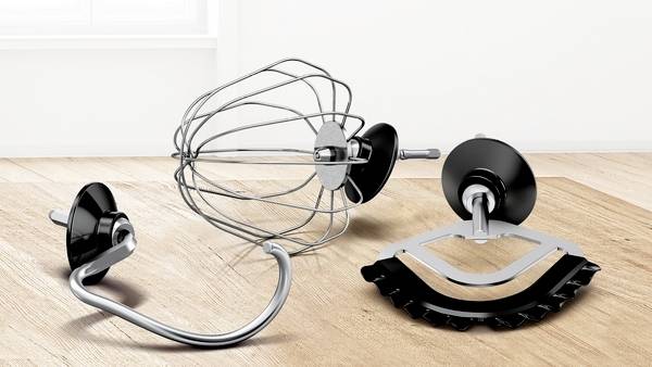 Drei Küchenmaschinenaufsätze, darunter ein Drahtschneebesen, ein Knethaken und ein Flachrührer, werden auf einer Holzoberfläche präsentiert.