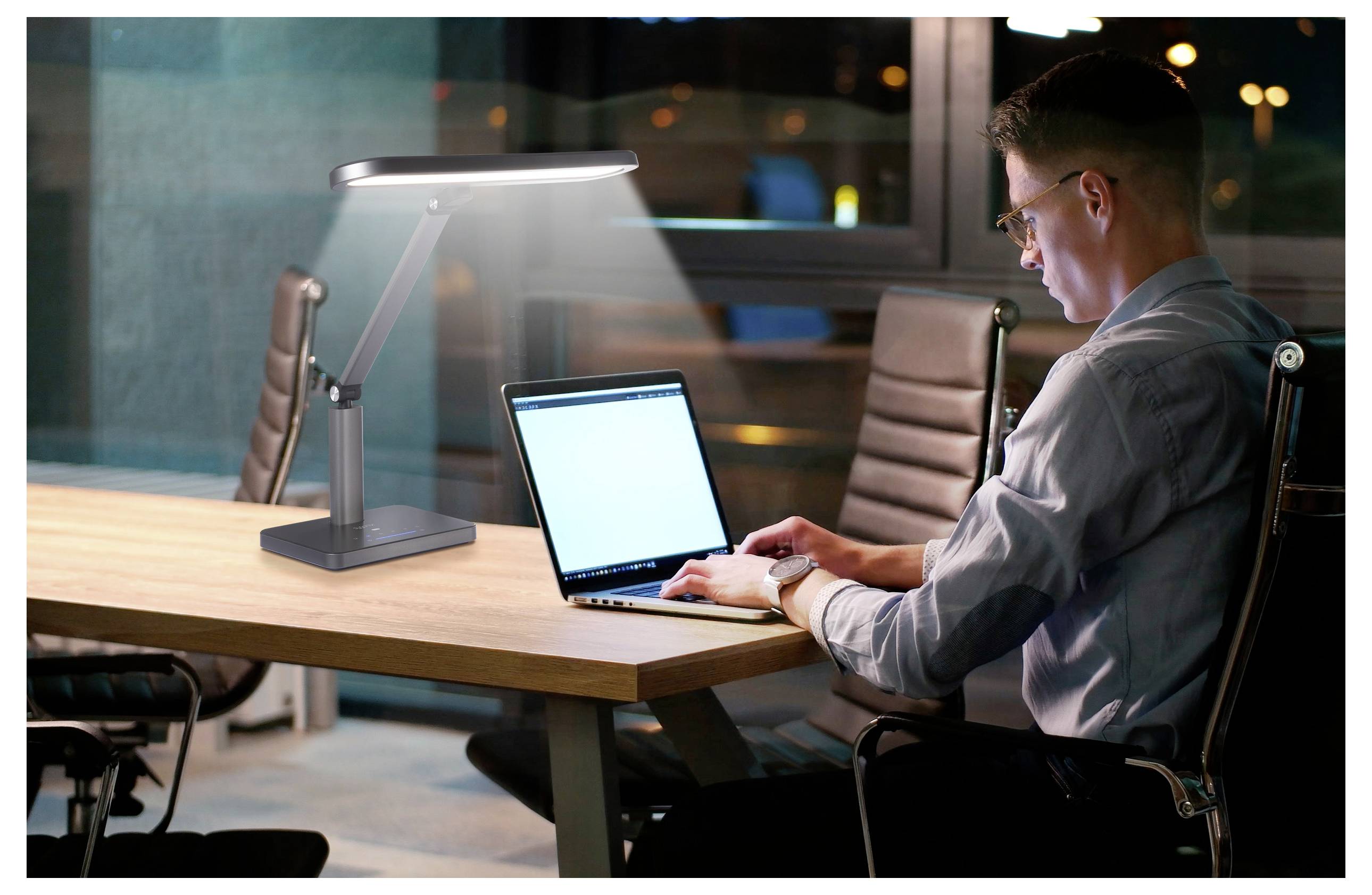 Ein Mann in einem Büro arbeitet an einem Laptop, beleuchtet von einer Schreibtischlampe, mit großen Fenstern und Stadtlichtern, die im Hintergrund zu sehen sind.