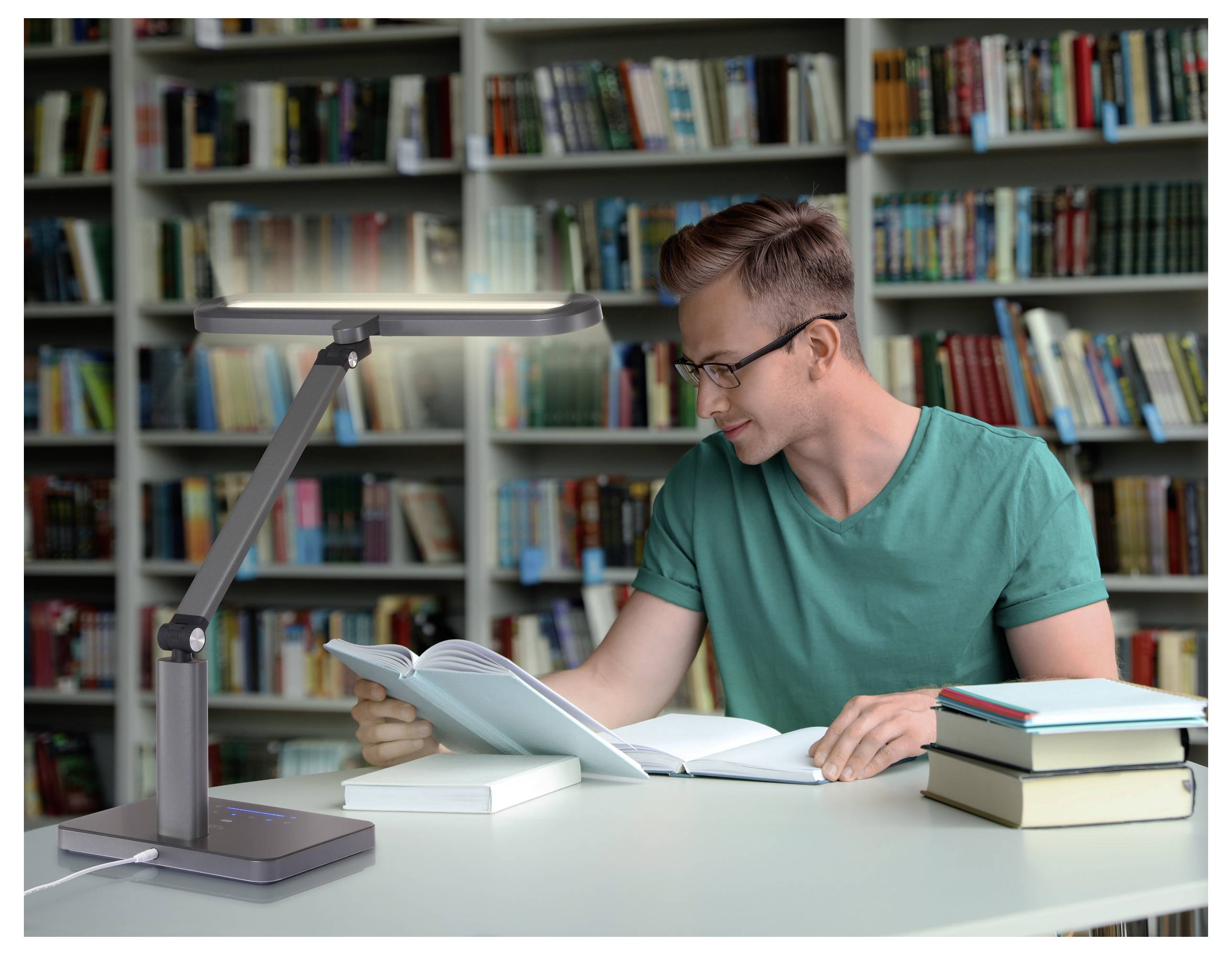 Eine Person sitzt an einem Bibliothekstisch und liest ein Buch unter einer modernen Schreibtischlampe. Im Hintergrund befinden sich Bücherregale, die mit Büchern gefüllt sind.