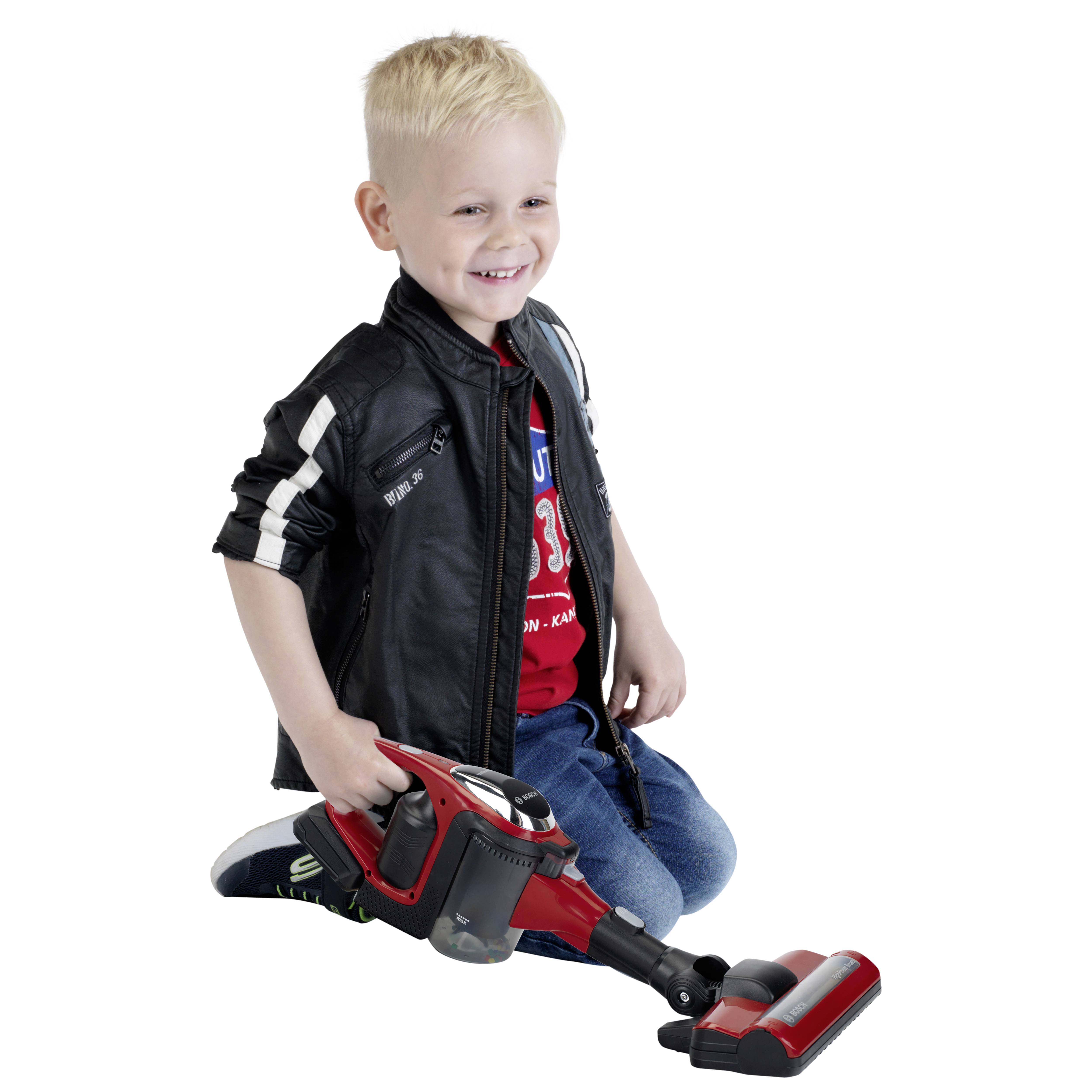 Ein kleines Kind mit blonder Frisur lächelt, während es auf dem Boden einen Spielzeug-Staubsauger hält und lässige Kleidung, einschließlich einer Jacke, trägt.