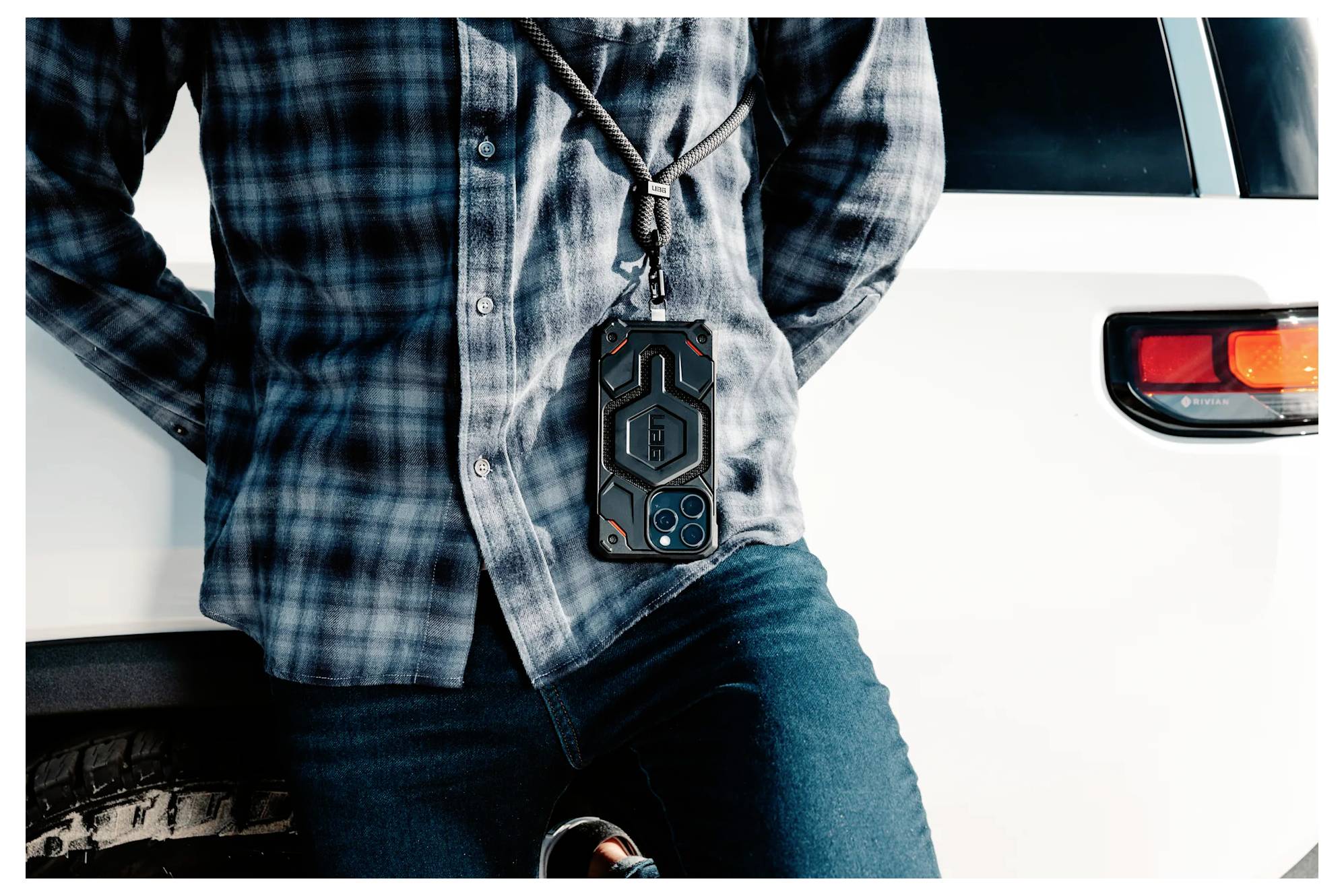 Eine Person in einem Karohemd lehnt an einem weißen SUV und trägt Jeans. An ihrem Hals hängt eine robuste, schwarze Handyhülle an einer Kordel.
