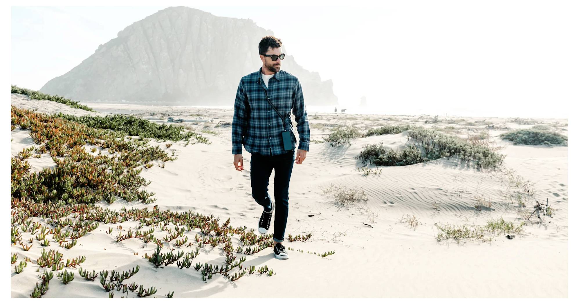 Eine Person mit Sonnenbrille läuft auf einem sandigen Strand mit spärlicher Vegetation und einer großen Felsformation im Hintergrund unter einem hellen Himmel.