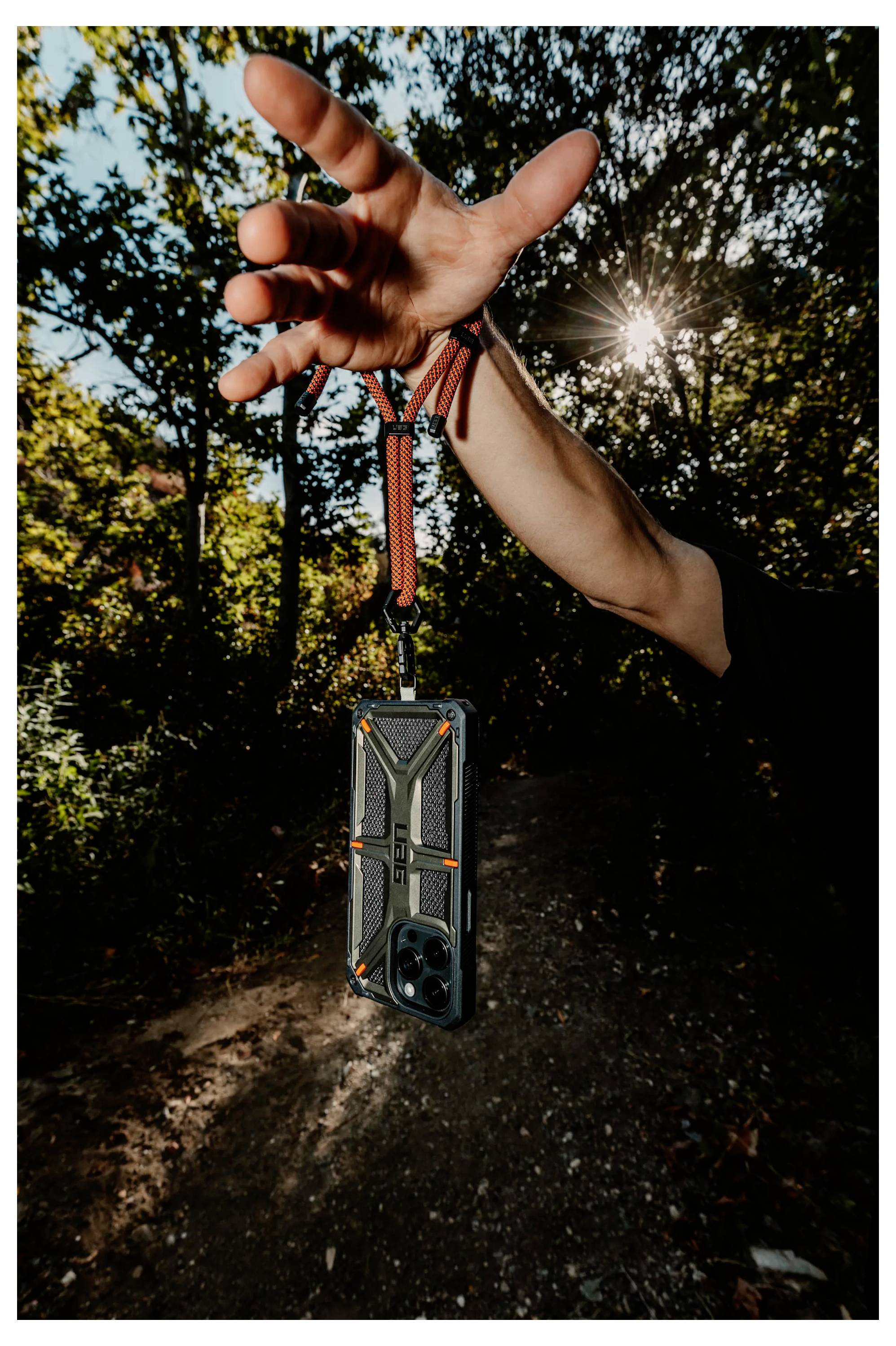 Eine Person hält ein Smartphone an einem Riemen in einem Wald aufgehängt, während Sonnenlicht durch die Bäume filtert. Die Szene deutet auf ein Outdoor-Abenteuer hin.