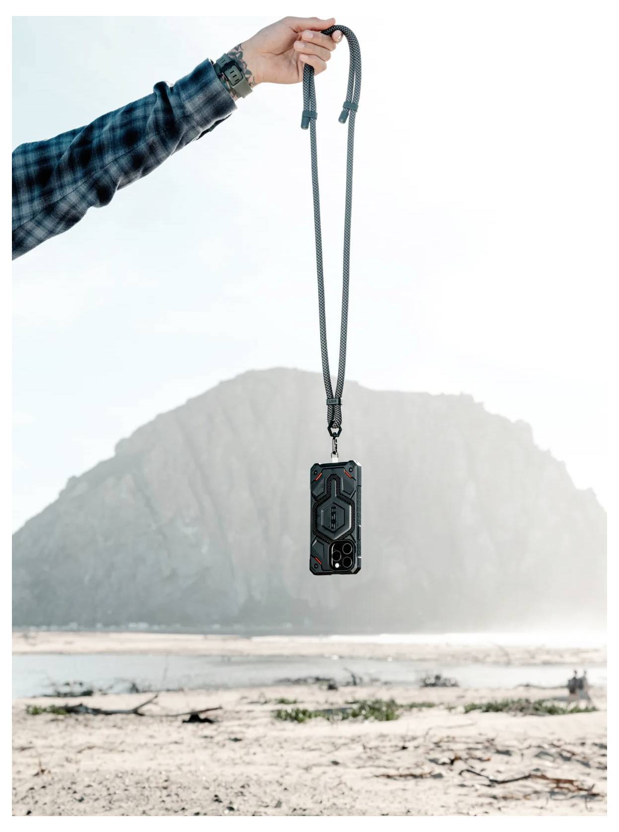 Eine Hand hält ein Handyhülle mit einem Trageriemen vor einer Strand-Kulisse, im Hintergrund eine große Felsformation.