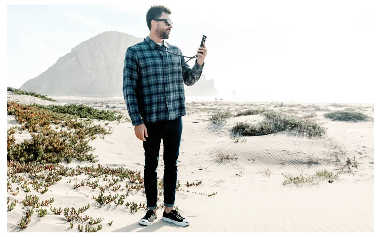Ein Mann mit Sonnenbrille steht auf einem sandigen Strand und hält ein Telefon. Im Hintergrund sind Vegetation und eine große Felsformation zu sehen.