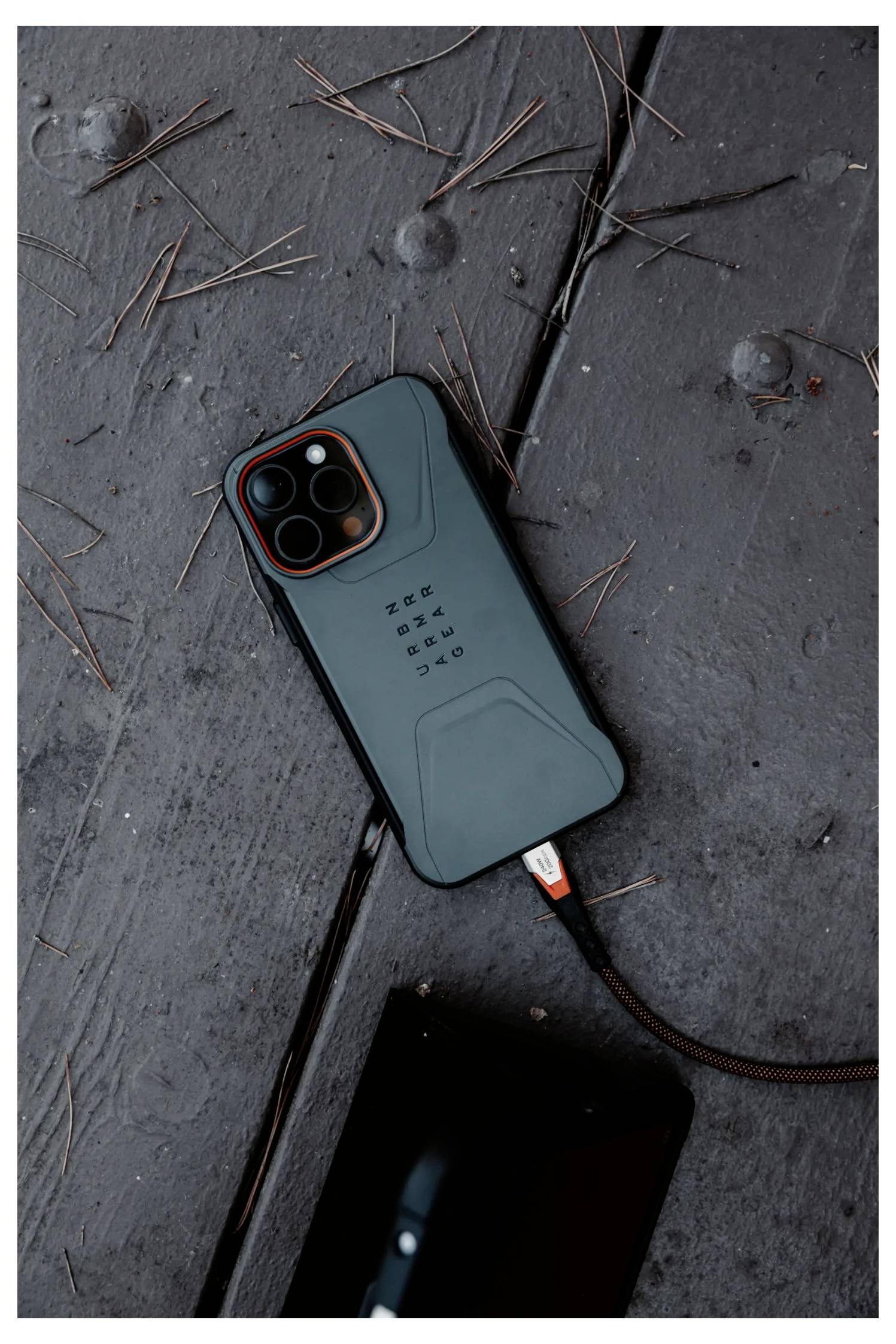 Ein Smartphone mit einer robusten Schutzhülle liegt auf einer Holzoberfläche und wird über ein sichtbares Kabel aufgeladen. Kiefernnadeln sind verstreut um das Gerät herum.