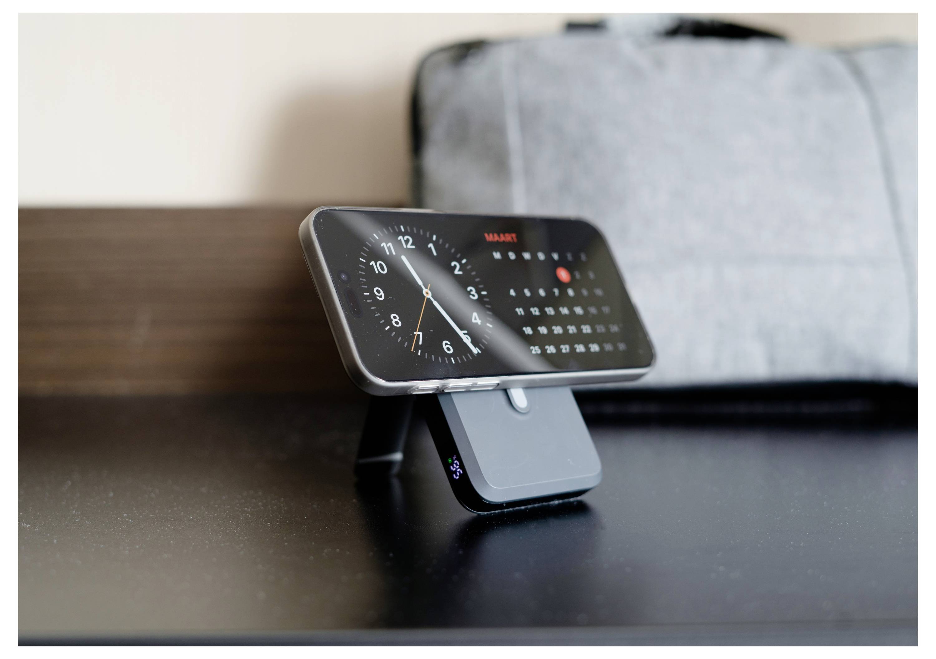 Ein Smartphone auf einem Ständer zeigt eine analoge Uhr und einen Kalender. Ein grauer Beutel ist im Hintergrund unscharf.