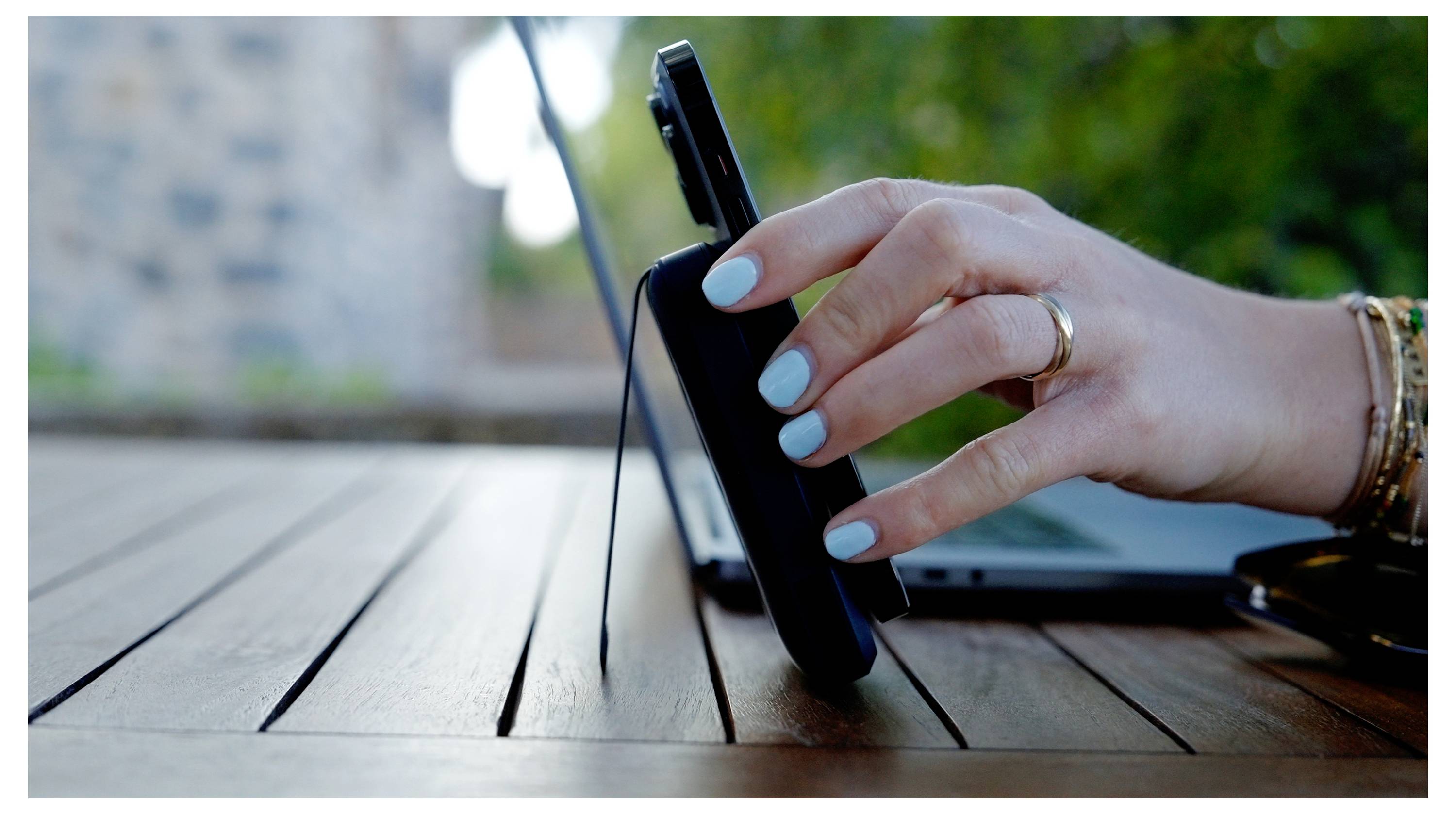 Eine Hand mit hellblauen Nägeln und Armbändern berührt ein Smartphone auf einem Holztisch im Freien, während ein Laptop im Hintergrund zu sehen ist.