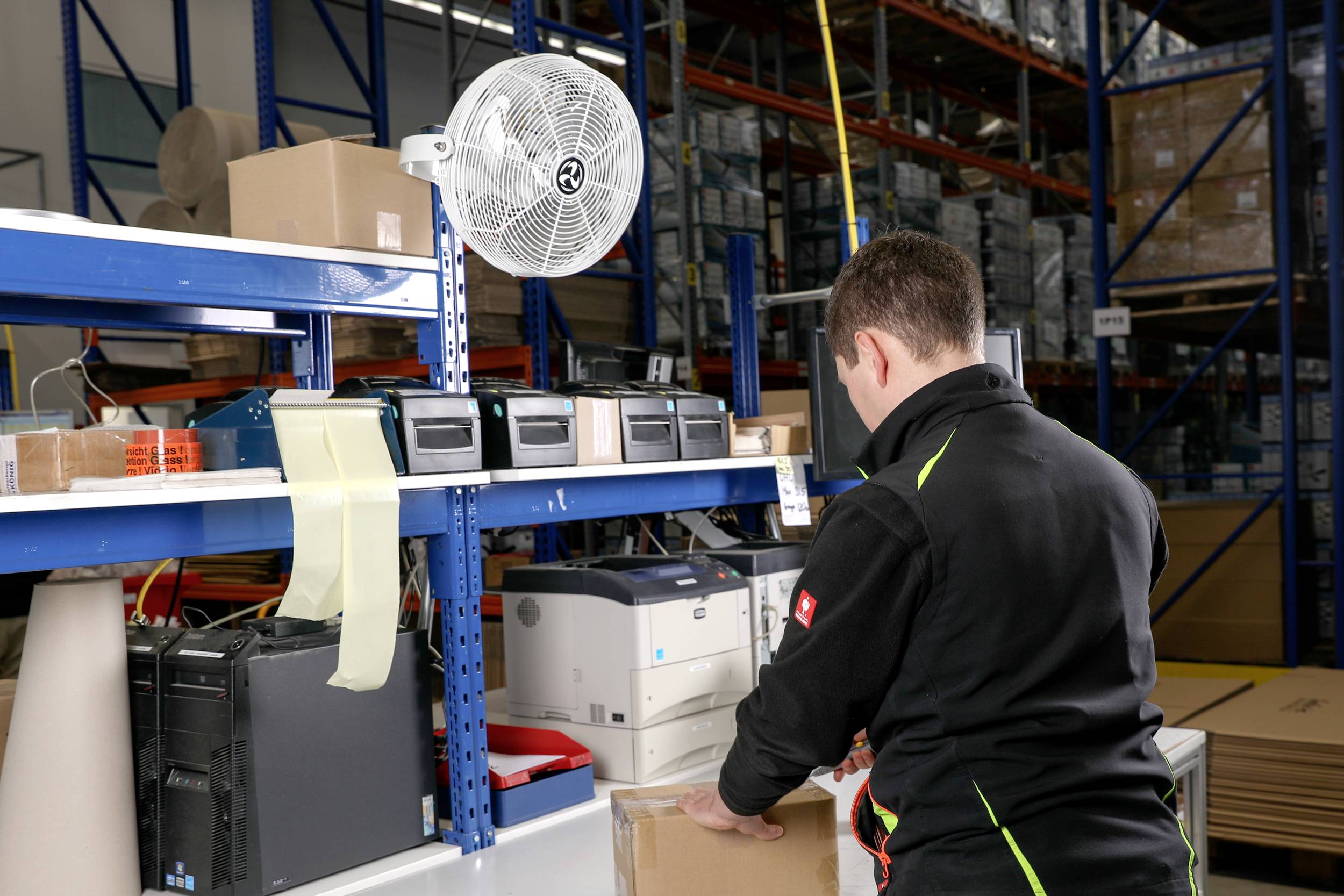 Eine Person verpackt eine Schachtel an einem Arbeitsplatz in einem Lager. Es gibt Kartons und elektronische Geräte in der Umgebung sowie Regale voller Inventar.