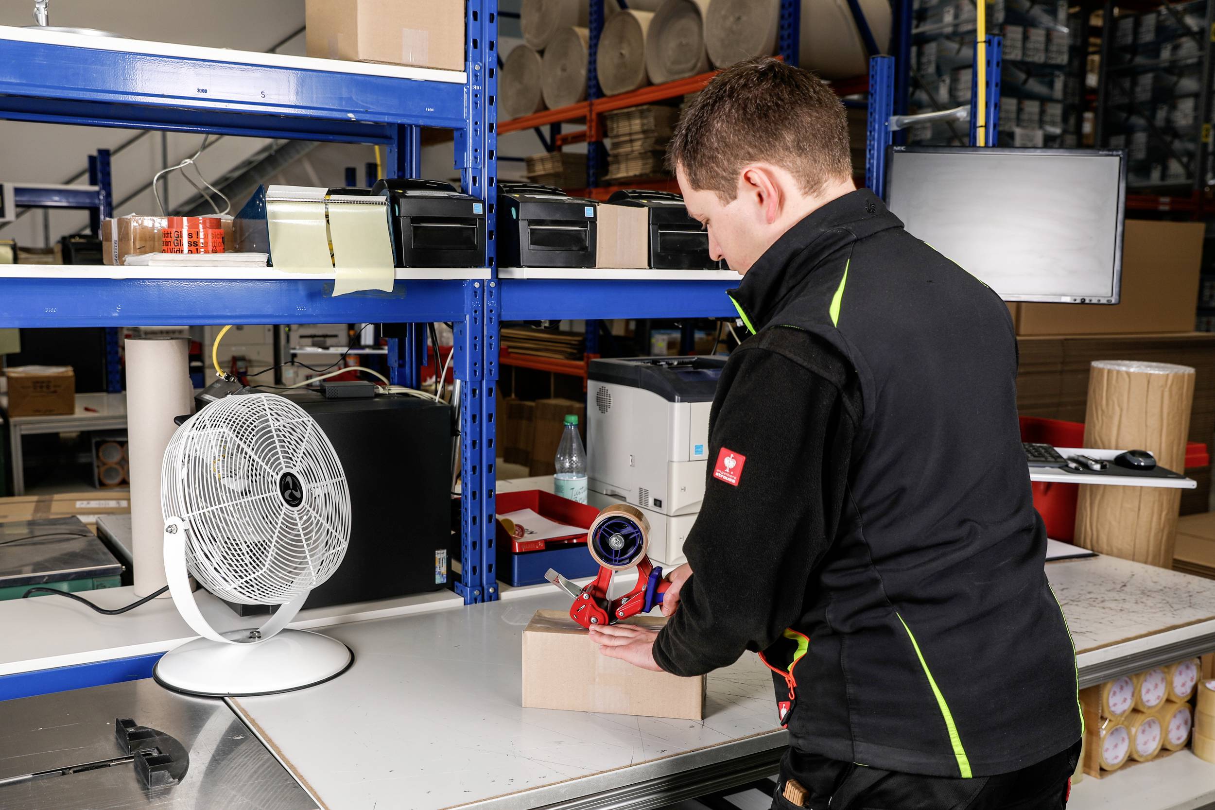 Eine Person in schwarzer Uniform verwendet Klebeband, um eine Schachtel an einem Arbeitsplatz mit Regalen, Ventilator und Verpackungsmaterialien in einem organisierten Arbeitsbereich zu verschließen.