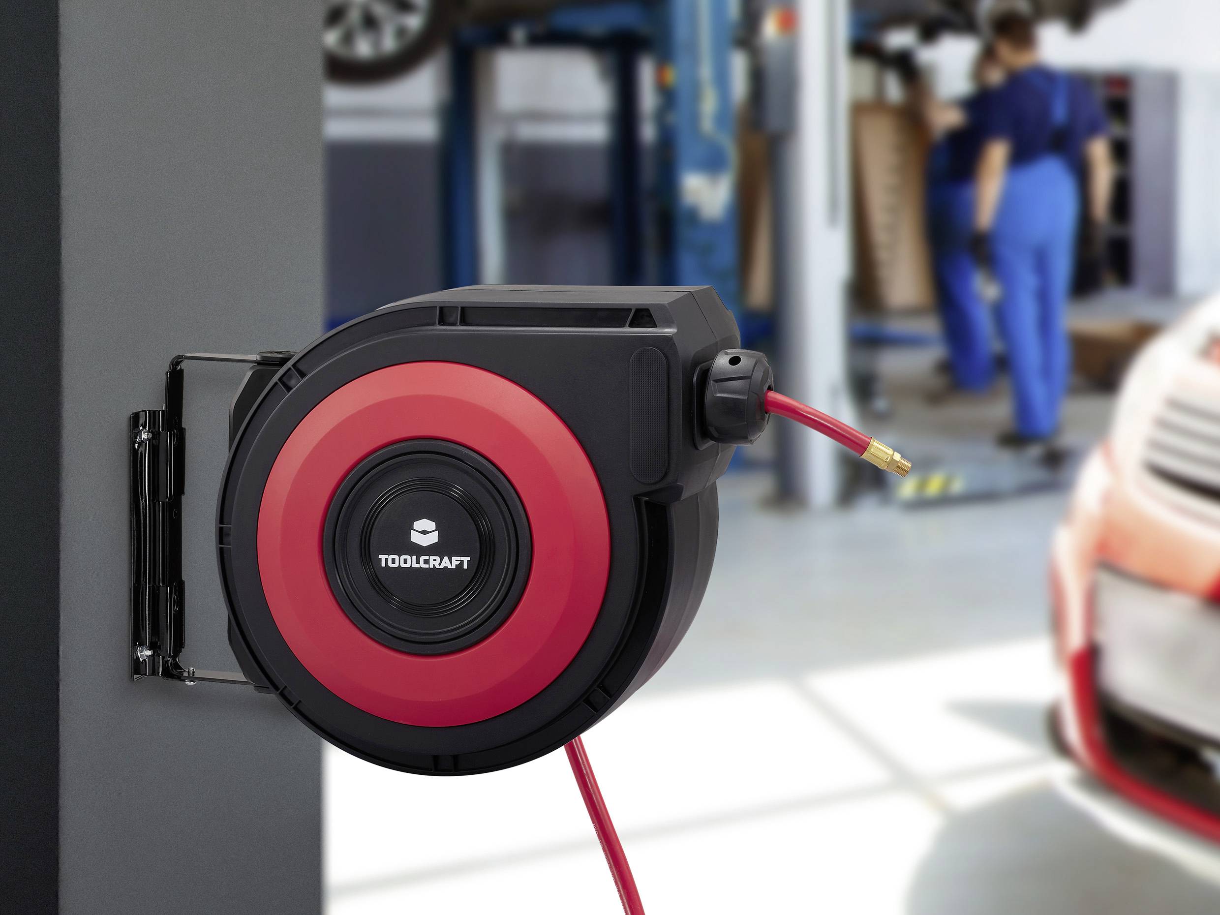 Rot-schwarze Toolcraft Schlauchtrommel, an einer Wand in einer Autowerkstatt montiert, mit einem Mechaniker, der im Hintergrund arbeitet.