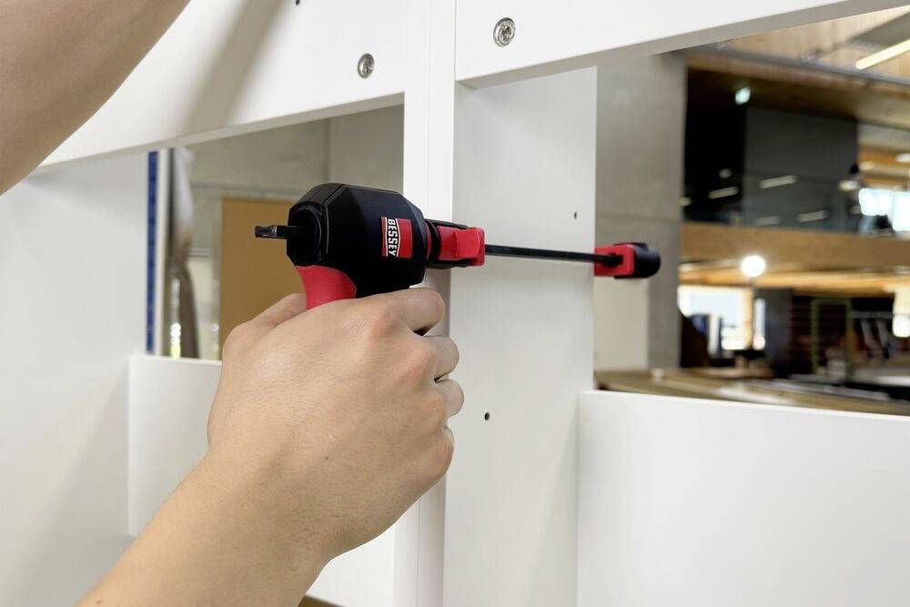 Eine Person verwendet eine schwarze und rote Bohrmaschine, um eine Schraube in ein weißes Holzbrett zu schrauben, wobei eine Büroumgebung im Hintergrund sichtbar ist.
