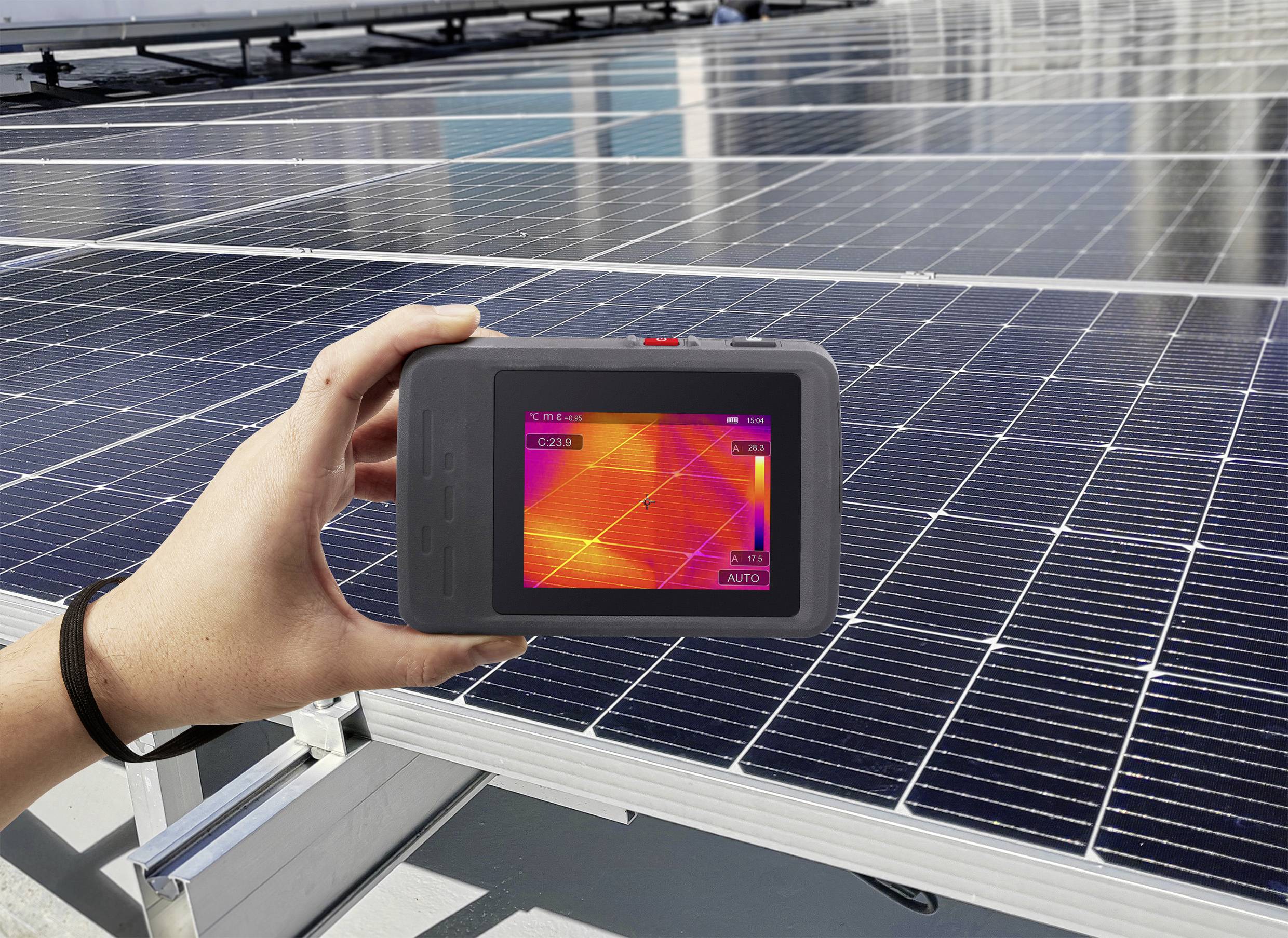 Eine Person hält ein Wärmebildgerät, das eine Wärmekarte von Solarpanels zeigt, während die Panels unter einem klaren Himmel zu sehen sind.