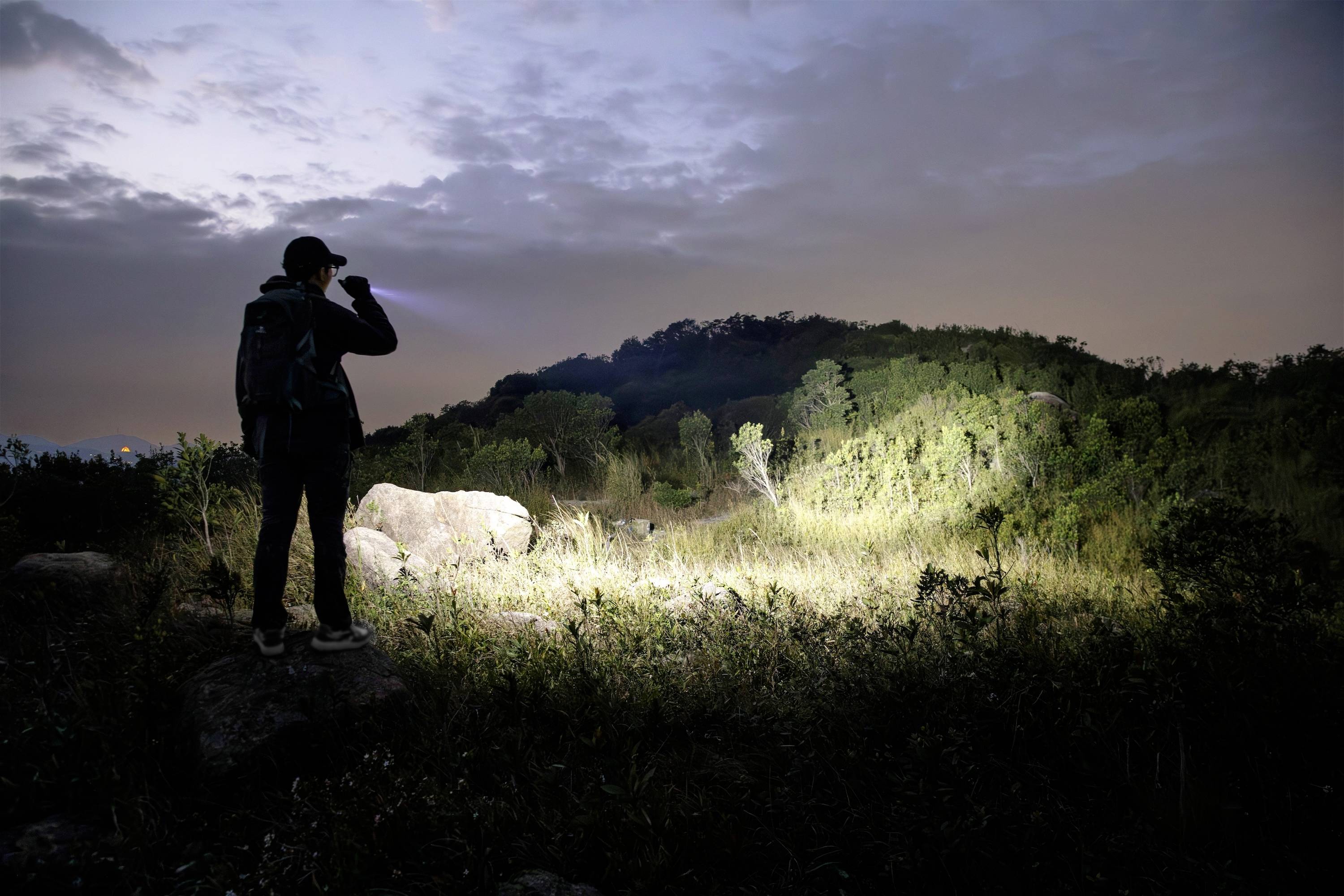 Eine Person mit einem Rucksack steht auf einem Stein in einem grasigen Gebiet bei Einbruch der Dunkelheit und leuchtet mit einer Taschenlampe auf einen Hügel mit spärlicher Vegetation.