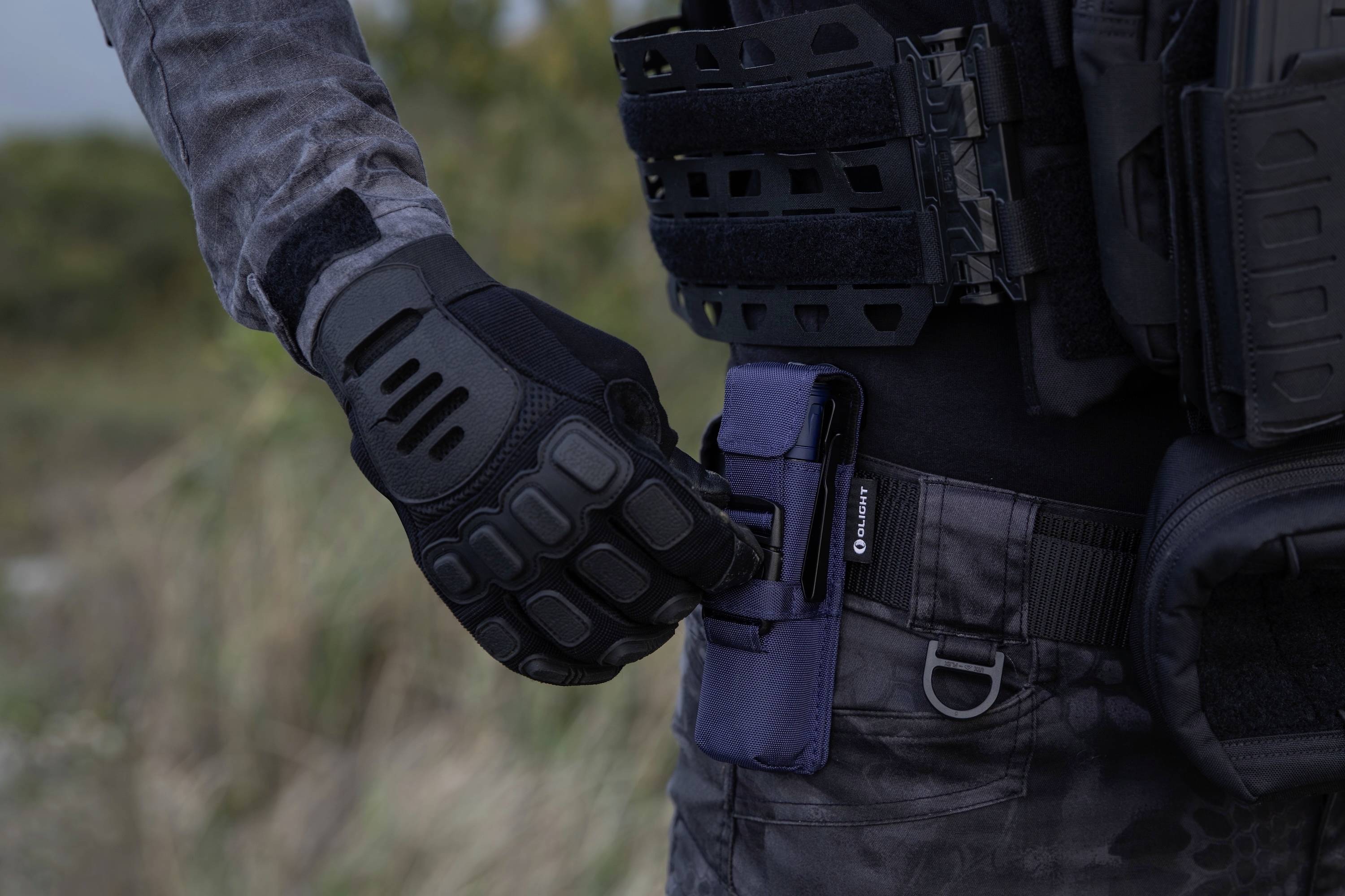 Eine Person in taktischer Ausrüstung und Handschuhen justiert ein Werkzeug oder Gerät an ihrem Gürtel in einer Outdoor-Umgebung.