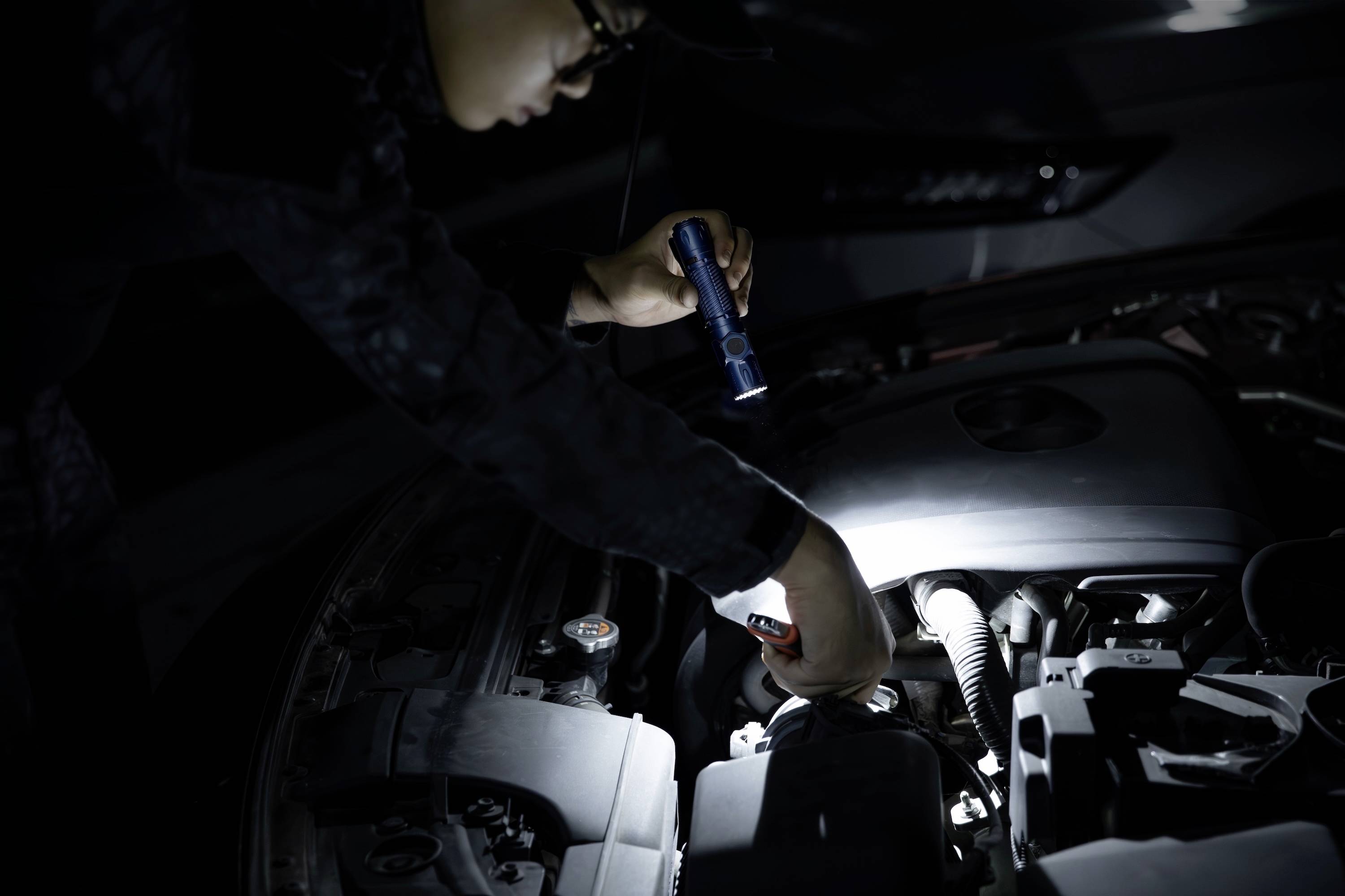 Eine Person untersucht einen Automotor mit einer Taschenlampe und deutet damit auf Wartungs- oder Fehlersuche in einer schlecht beleuchteten Umgebung hin.