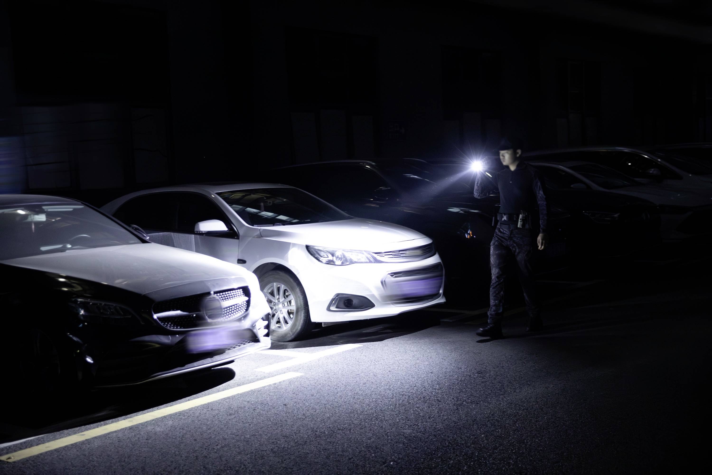 Ein Sicherheitswächter untersucht eine Reihe geparkter Autos auf einem schwach beleuchteten Parkplatz und leuchtet mit einer Taschenlampe bei der genauen Inspektion.