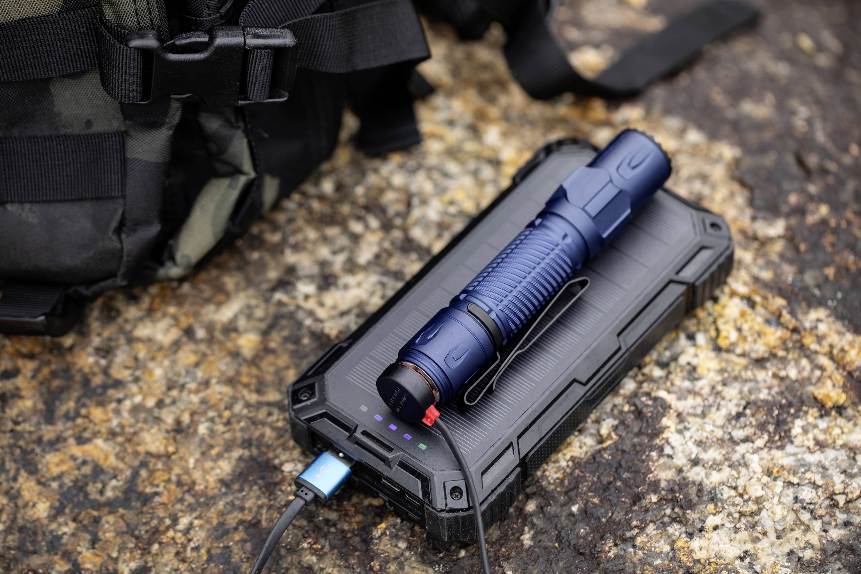 Eine blaue Taschenlampe liegt auf einem robusten schwarzen Powerbank, die über ein USB-Kabel verbunden ist und auf strukturiertem Stein neben einem Rucksack platziert ist.