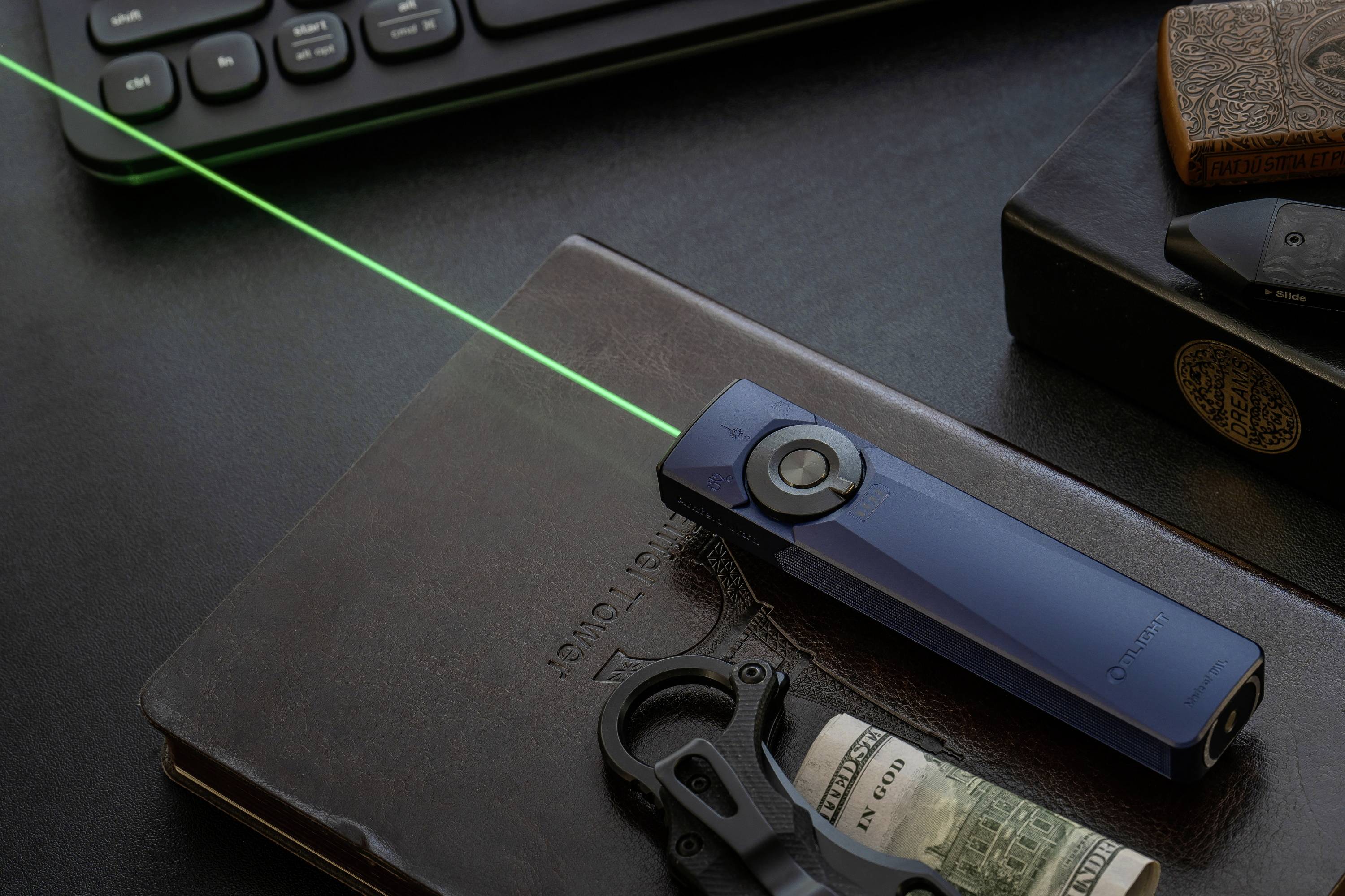 Ein blauer Laserpointer projiziert einen grünen Strahl auf einen Schreibtisch mit geschlossenen Büchern, einer Tastatur, einem Multitool und einer Geldrolle in der Nähe.