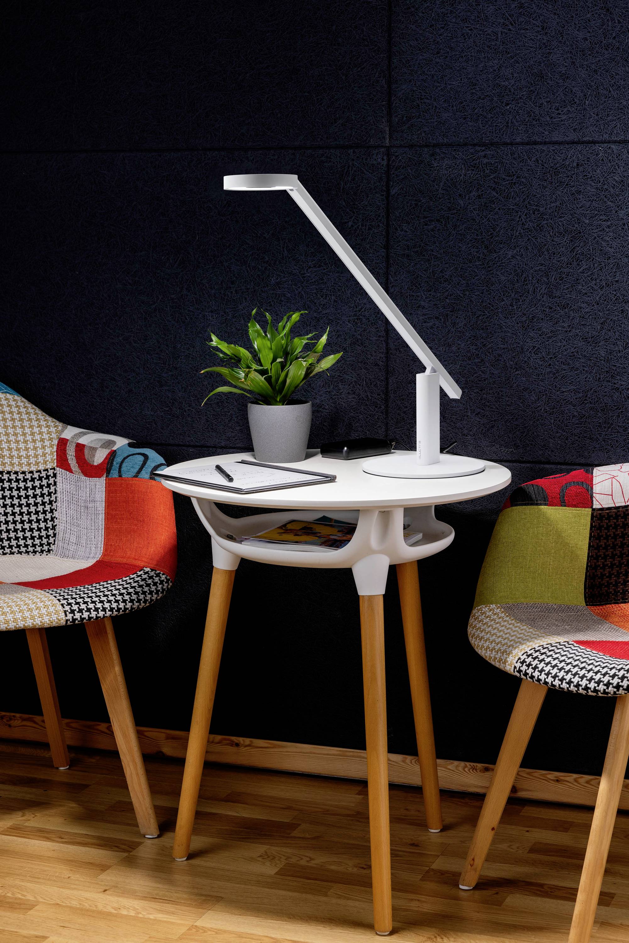 Ein kleiner weißer Tisch mit einer modernen Lampe, einer Topfpflanze sowie einem Stift und einem Notizbuch, flankiert von zwei farbenfrohen, gemusterten Stühlen.