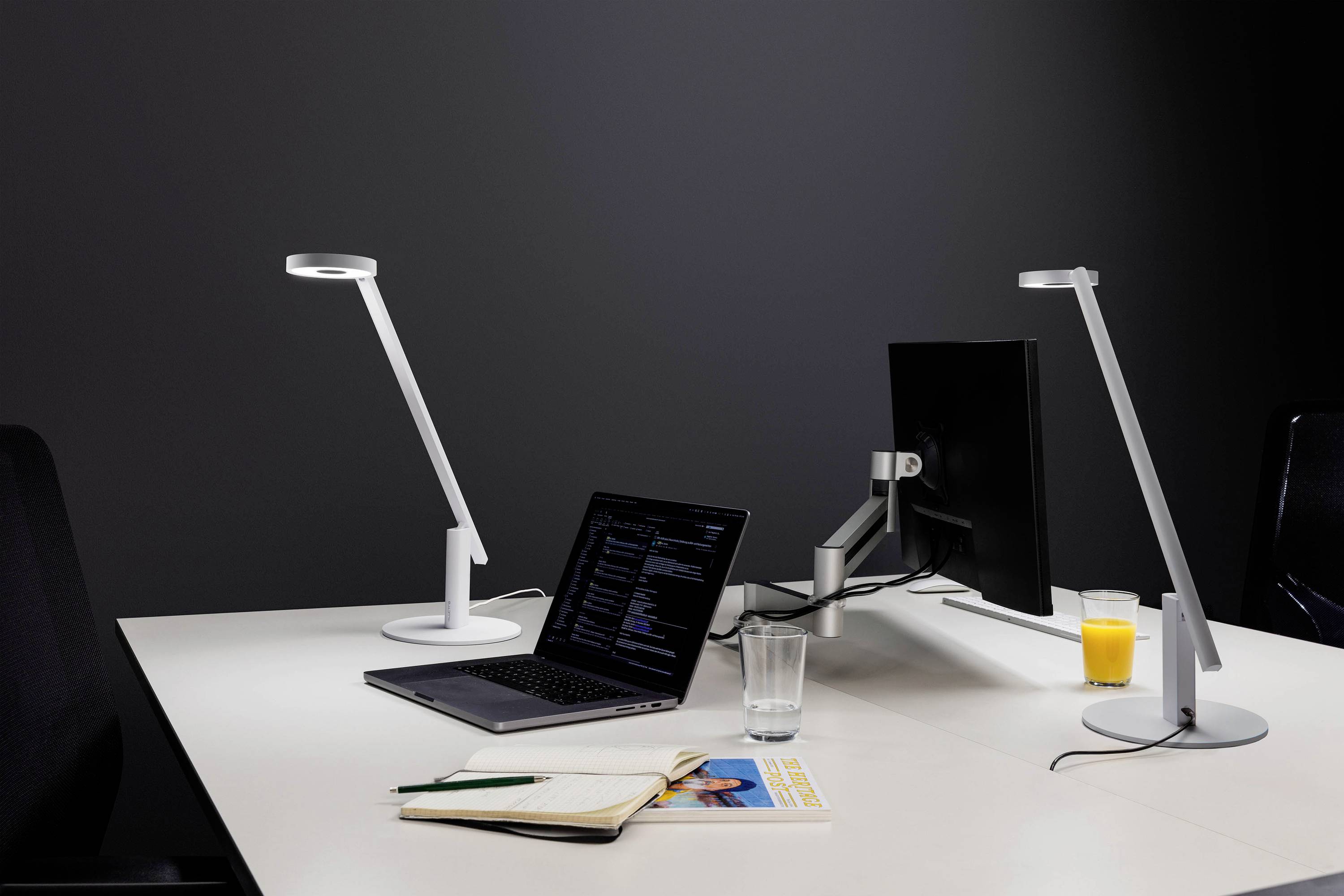 Ein moderner Arbeitsplatz mit einem Laptop, auf dem Code angezeigt wird, einem Monitor, Schreibtischlampen, einem aufgeschlagenen Notizbuch, einem Glas Wasser und einem Glas Orangensaft.