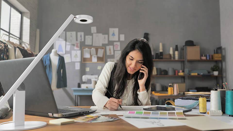 Eine Frau in einem Designstudio telefoniert, während sie an einem Schreibtisch skizziert. Stoffe, ein Laptop und Designmaterialien sind zu sehen.