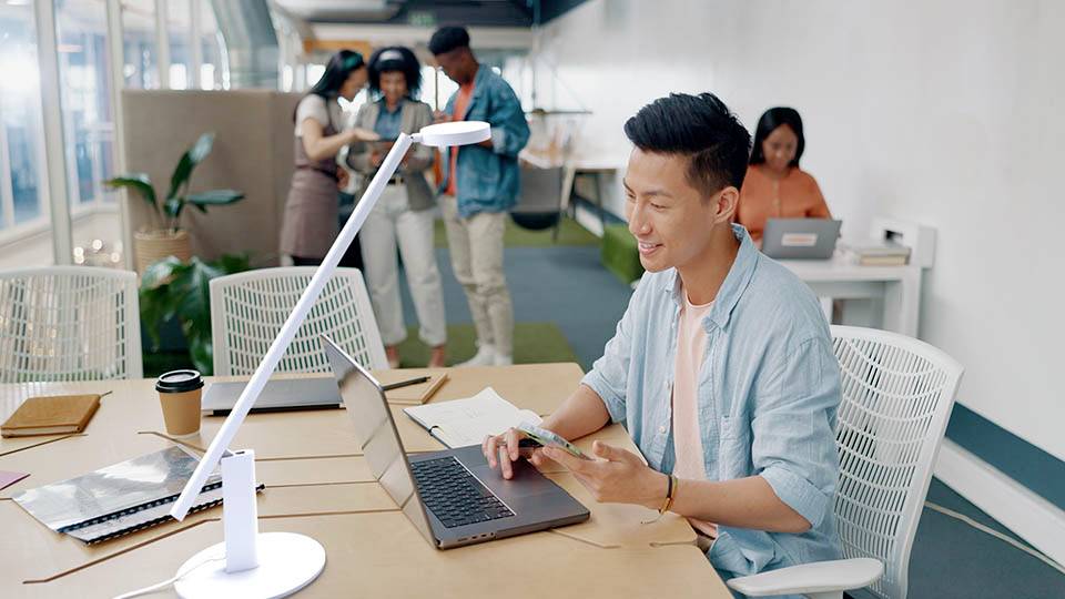 Eine Person, die an einem Laptop in einem hellen, modernen Büro arbeitet und ein Smartphone in der Hand hält, während im Hintergrund drei Kollegen Papiere besprechen.
